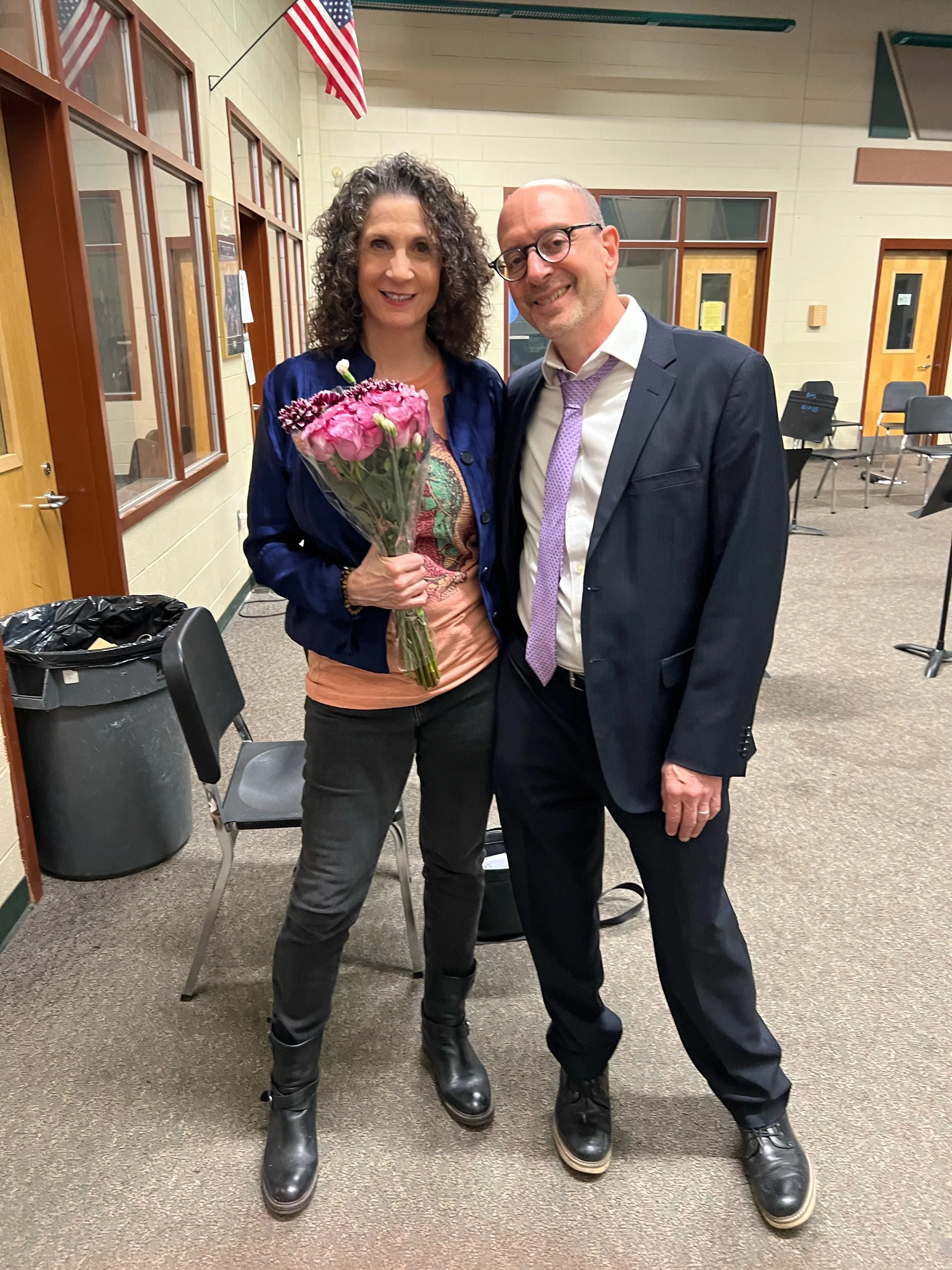 A woman holding a bouquet of pink flowers standing next to a man in a suit inside a room with beige walls, windows, and American flags, with chairs and a trash bin in the background.