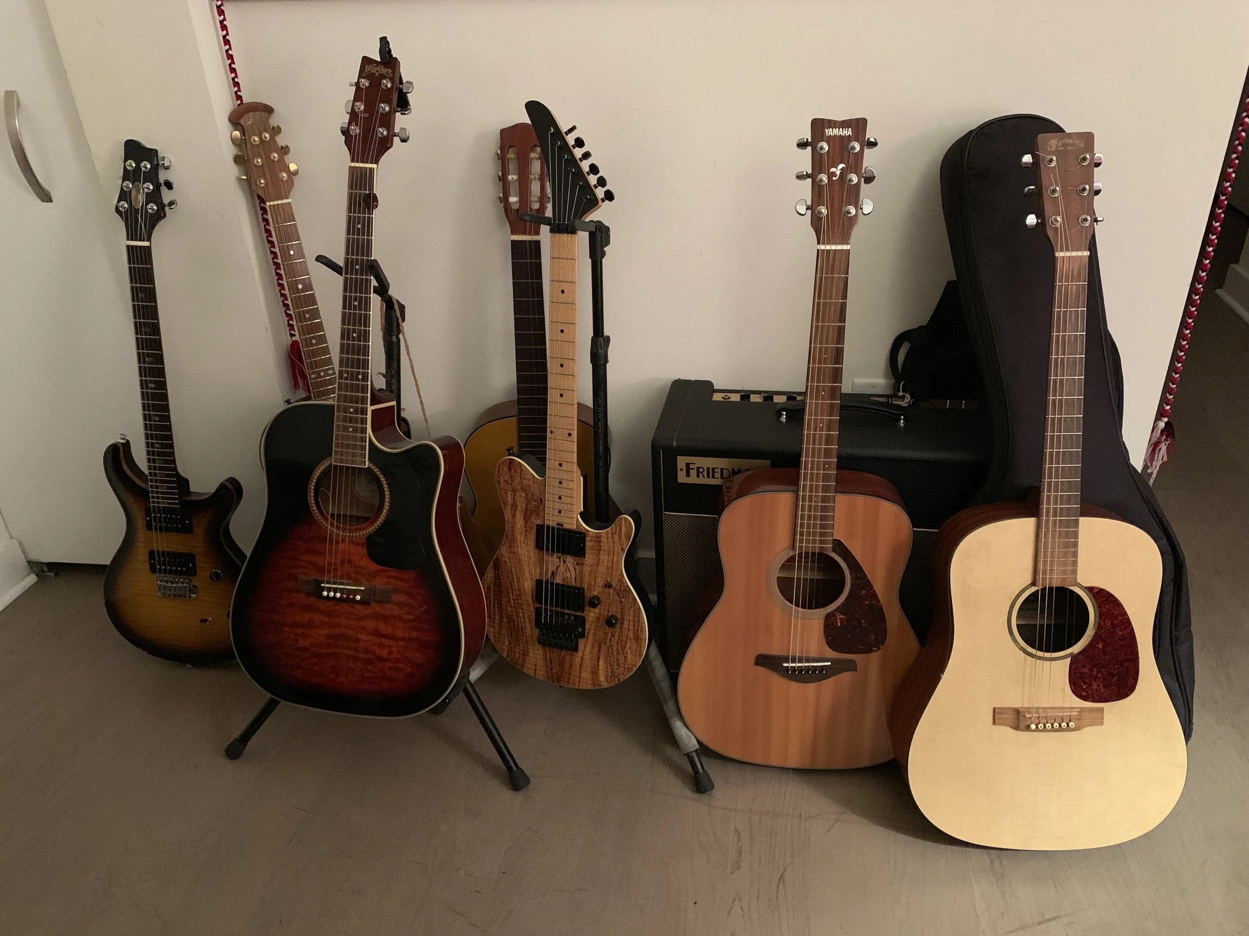 Five guitars of various styles and sizes, including an electric guitar, acoustic guitars, and a bass guitar, are arranged against a white wall, with a guitar case and amplifier nearby.