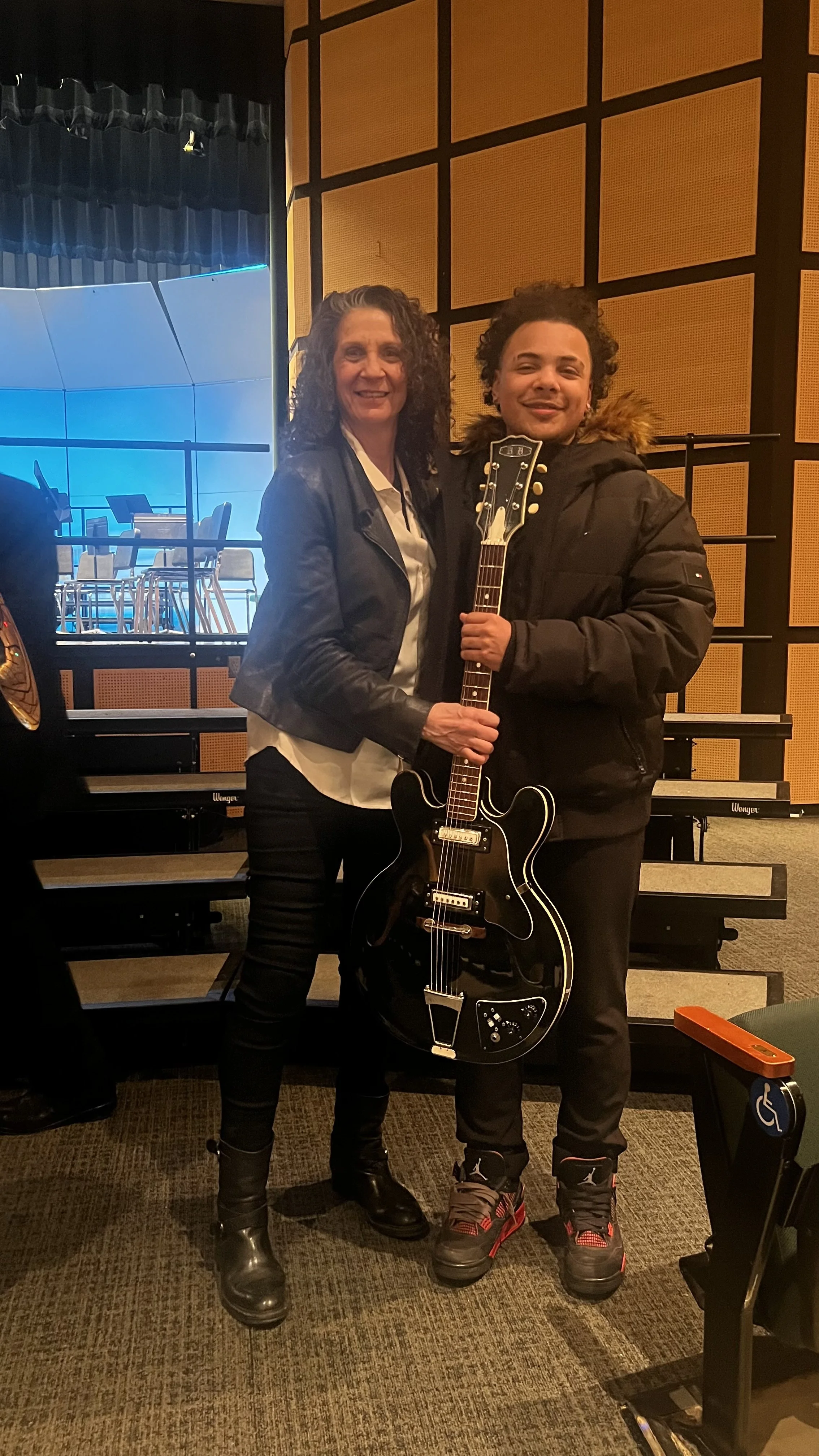 A woman and a young man holding a black electric guitar in a music rehearsal space or studio with a stage and chairs in the background.