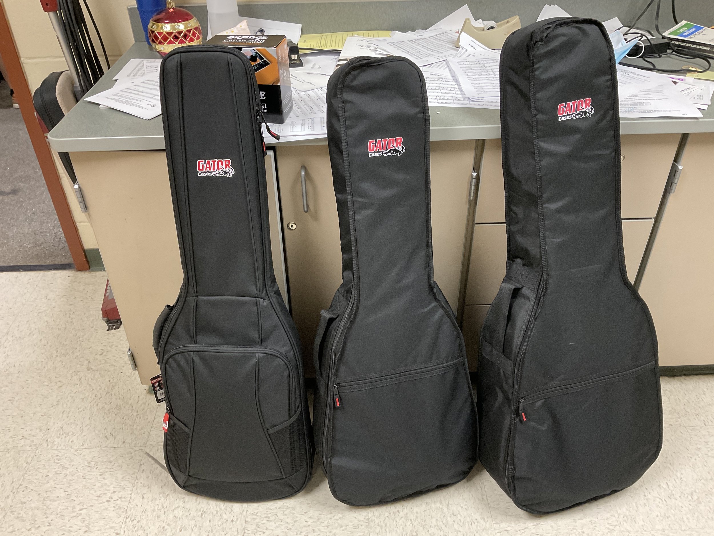 Three black guitar cases with GATOR Cases logo, standing upright next to a beige cabinet, in a room with papers and electronics on the counter.