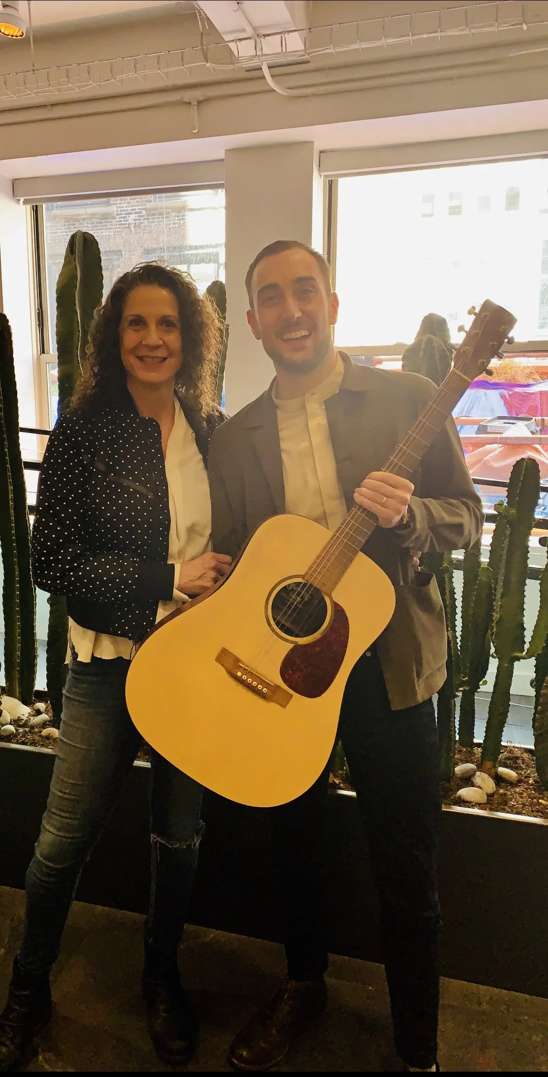 A woman with curly hair and a man with short hair smiling indoors, the man holding an acoustic guitar, with cactus plants and windows in the background.