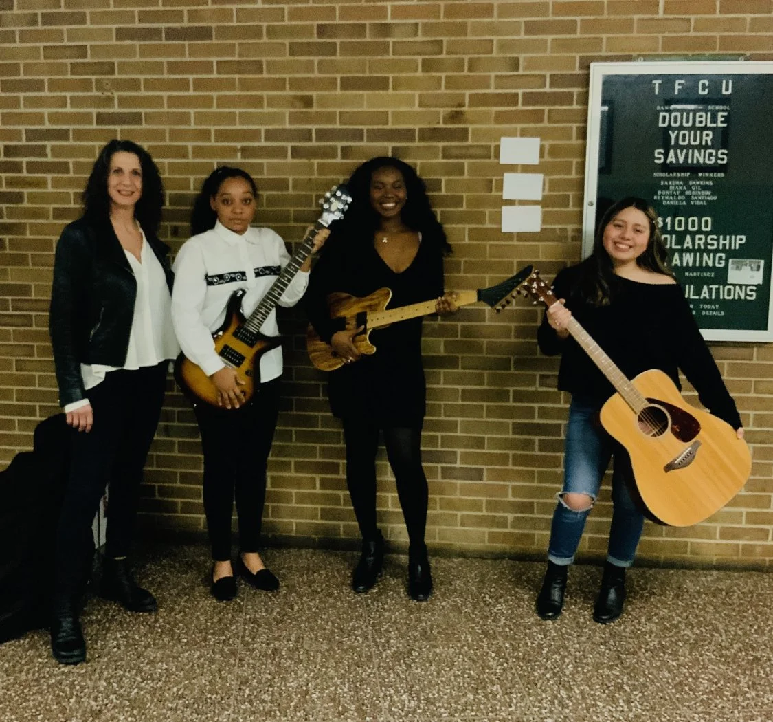 Four women standing against a brick wall, holding musical instruments including guitars, with a green sign on the wall.