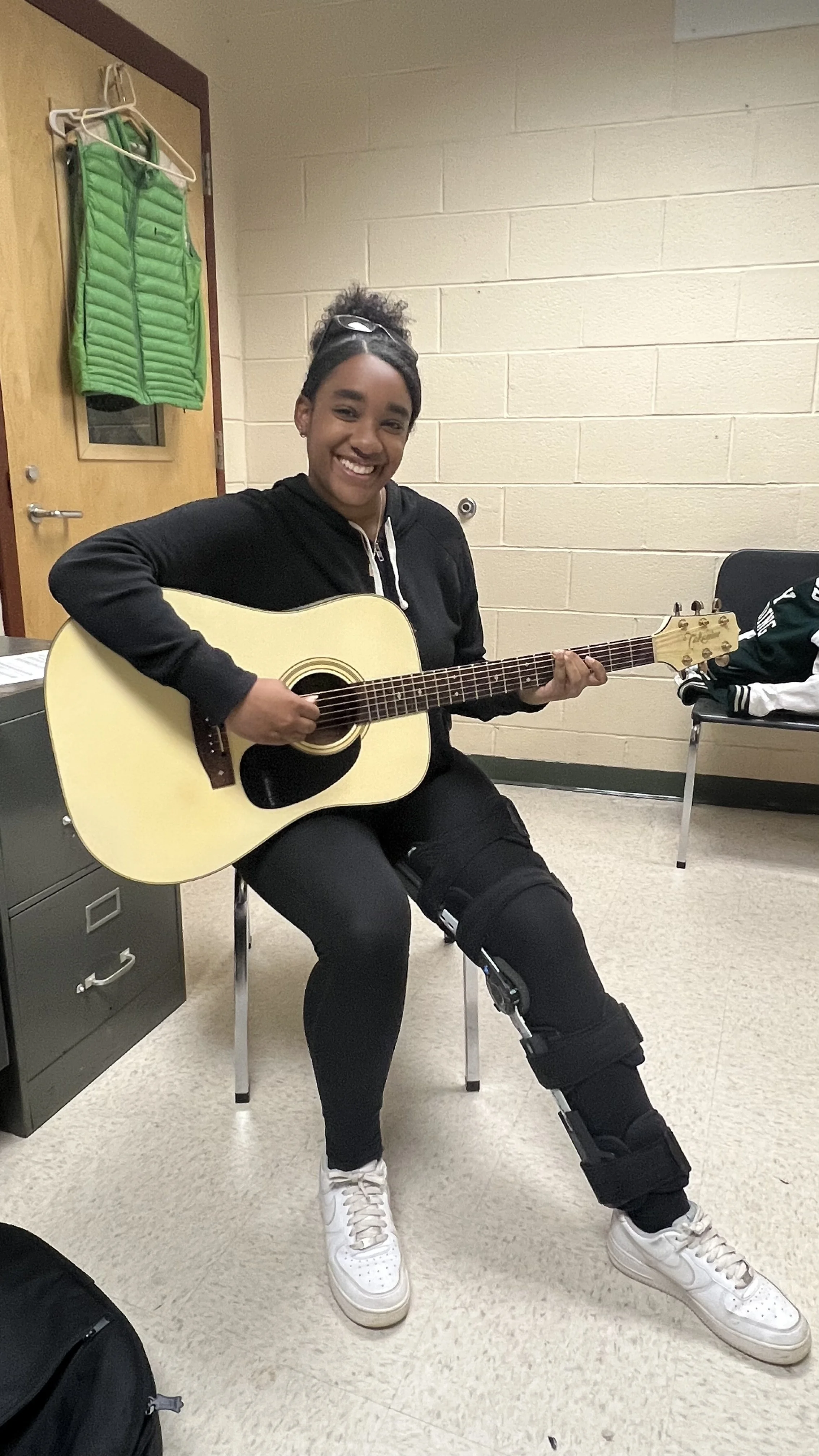 A young woman sitting on a chair, playing an acoustic guitar, smiling, in a room with beige cinder block walls, with a green vest hanging on the door.