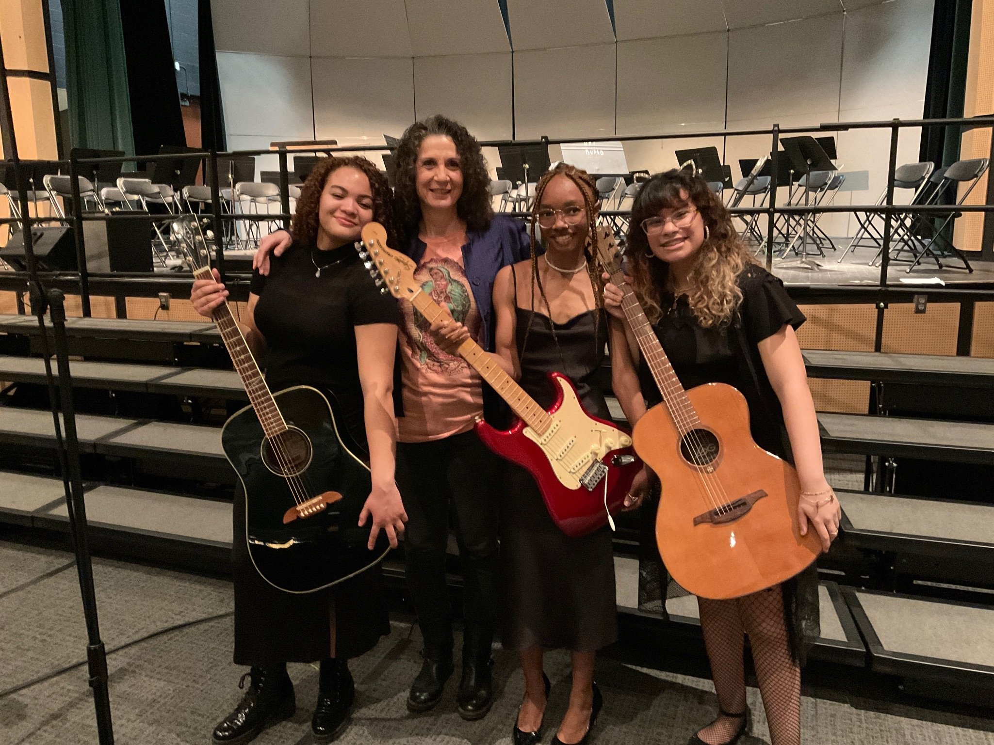 Four women standing on a stage holding guitars, with empty chairs and music stands behind them.