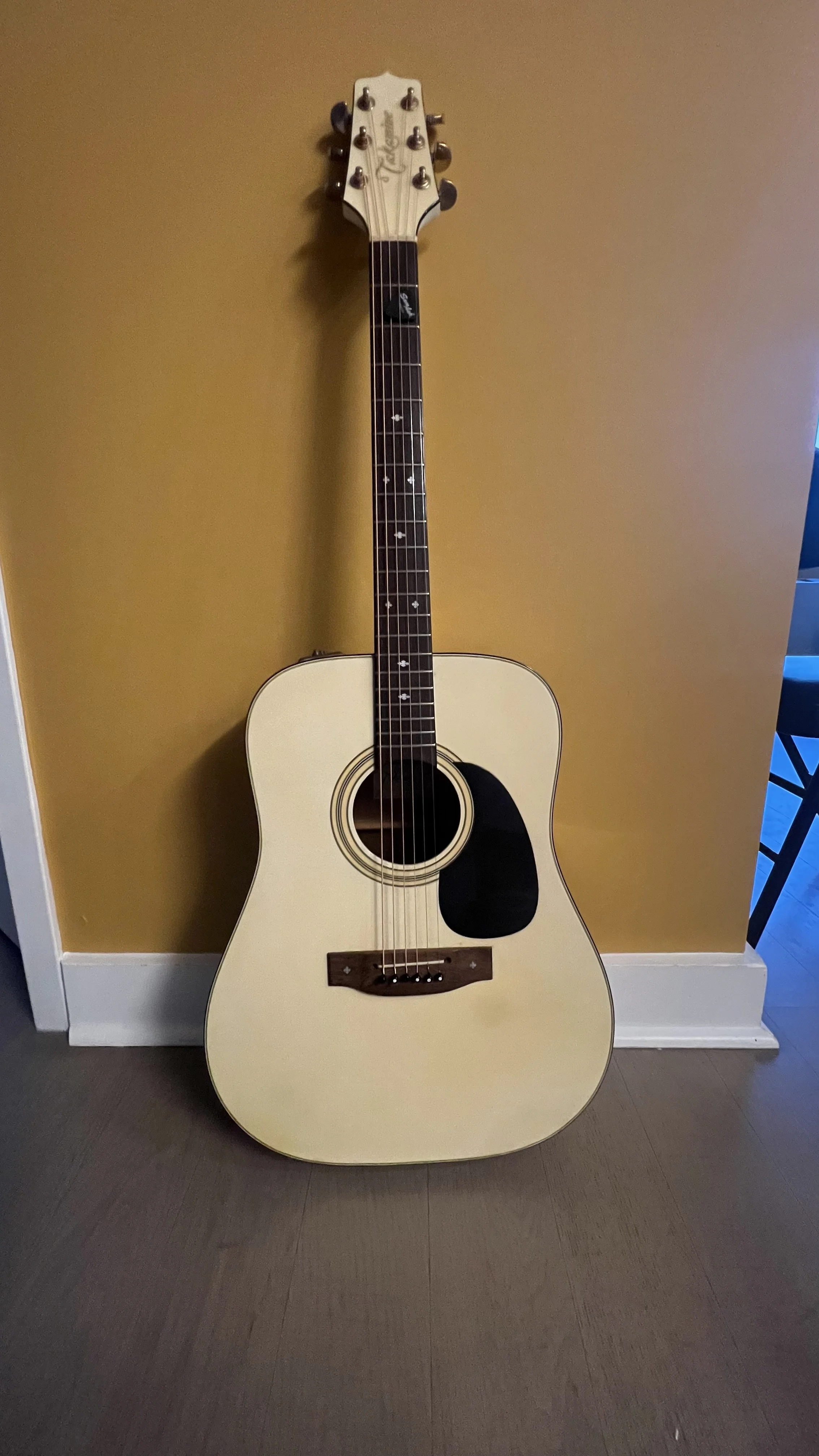 An acoustic guitar with a light-colored body, dark fretboard, and six strings, leaning against a yellow wall.