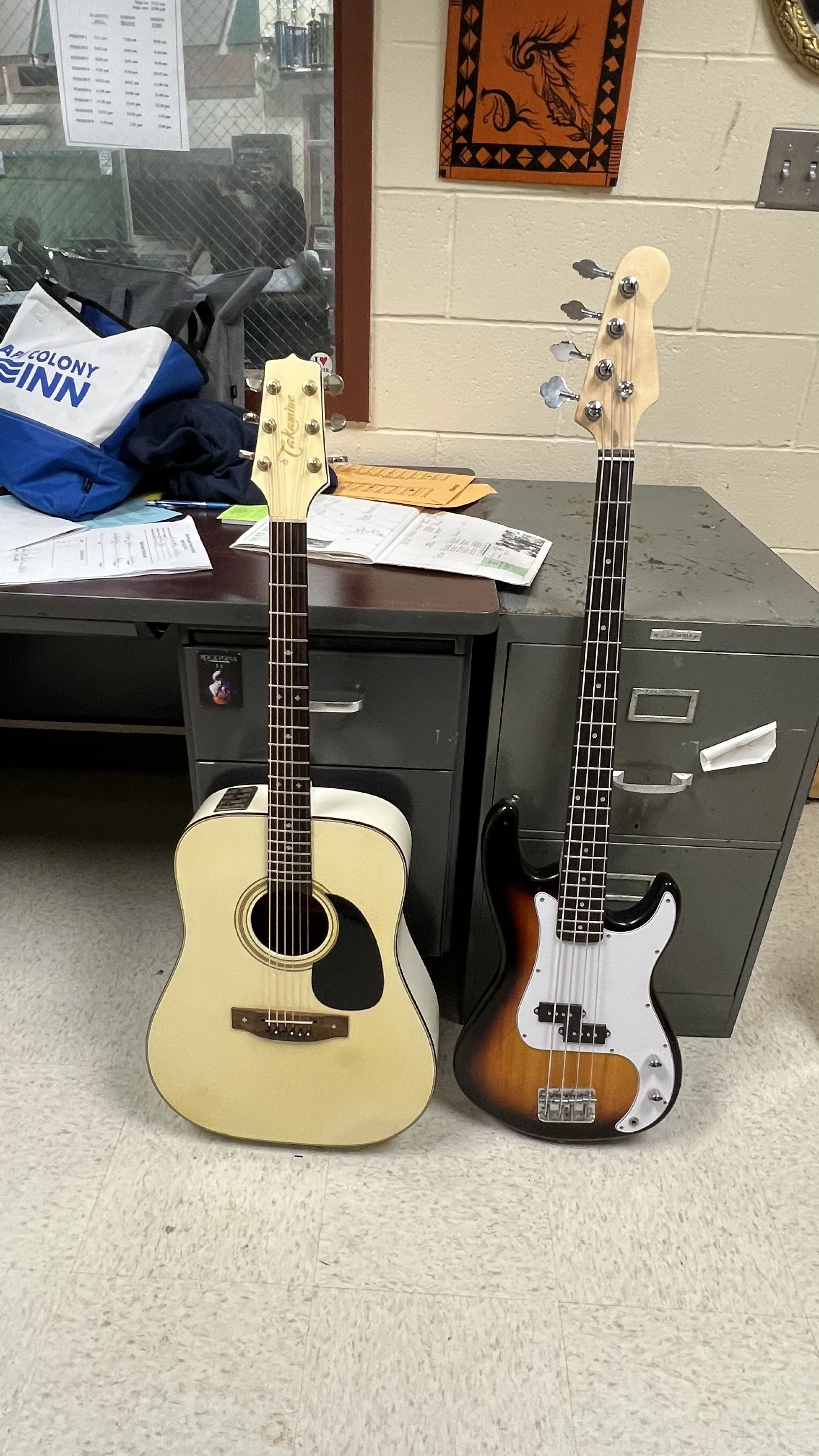 An acoustic guitar and a bass guitar leaning against a desk in a room, with papers and a bag on the desk, a window and a colorful wall hanging behind.