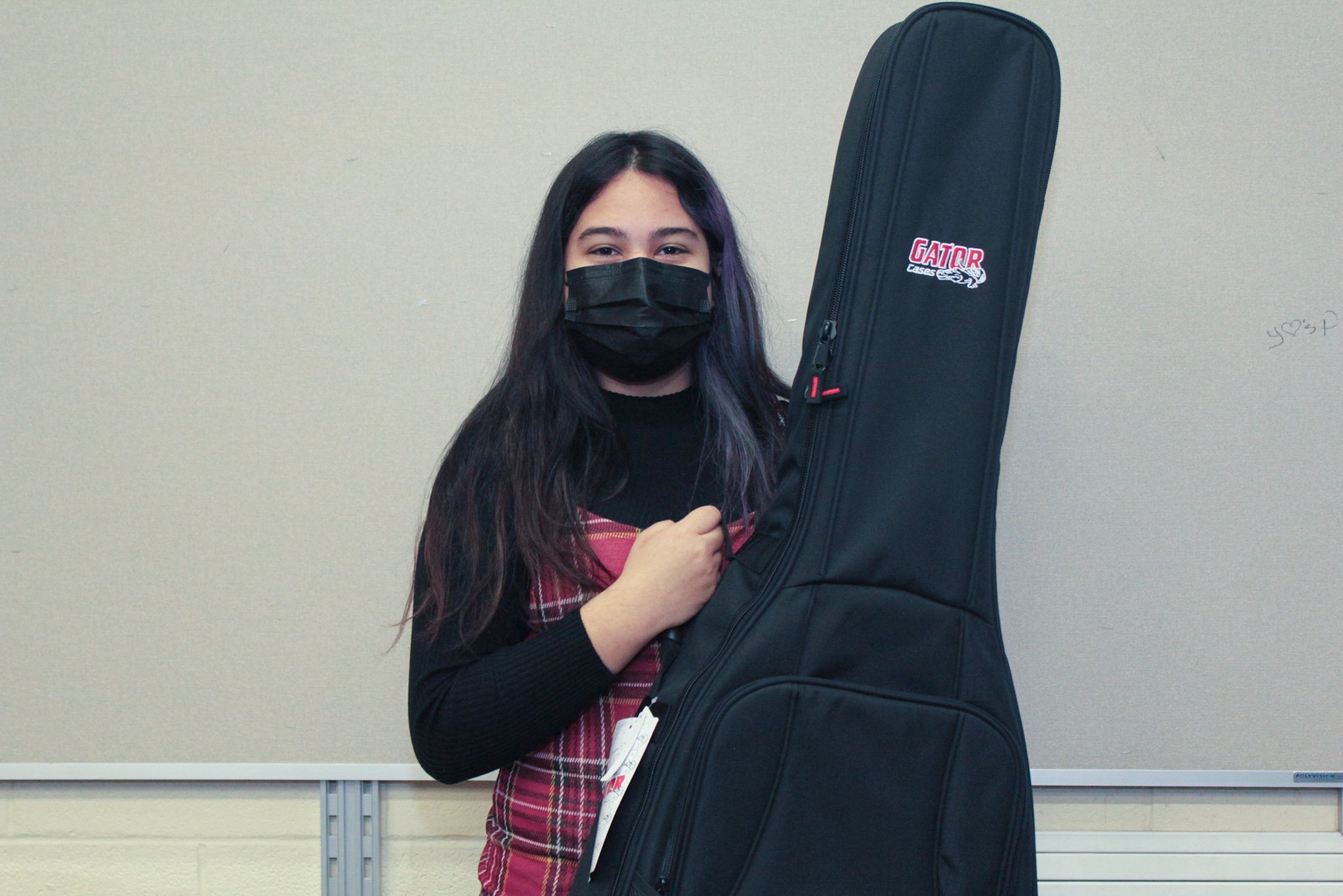 A young girl with long dark hair wearing a black face mask and a red plaid vest over a black long sleeve shirt, holding a black guitar case with a Gator logo, standing against a beige wall.