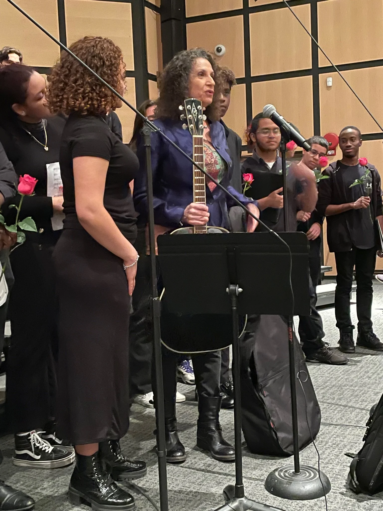 Group of people standing on stage during a musical or speech event, with a woman holding a guitar at the microphone, some holding roses, in an indoor setting with wood panel walls.