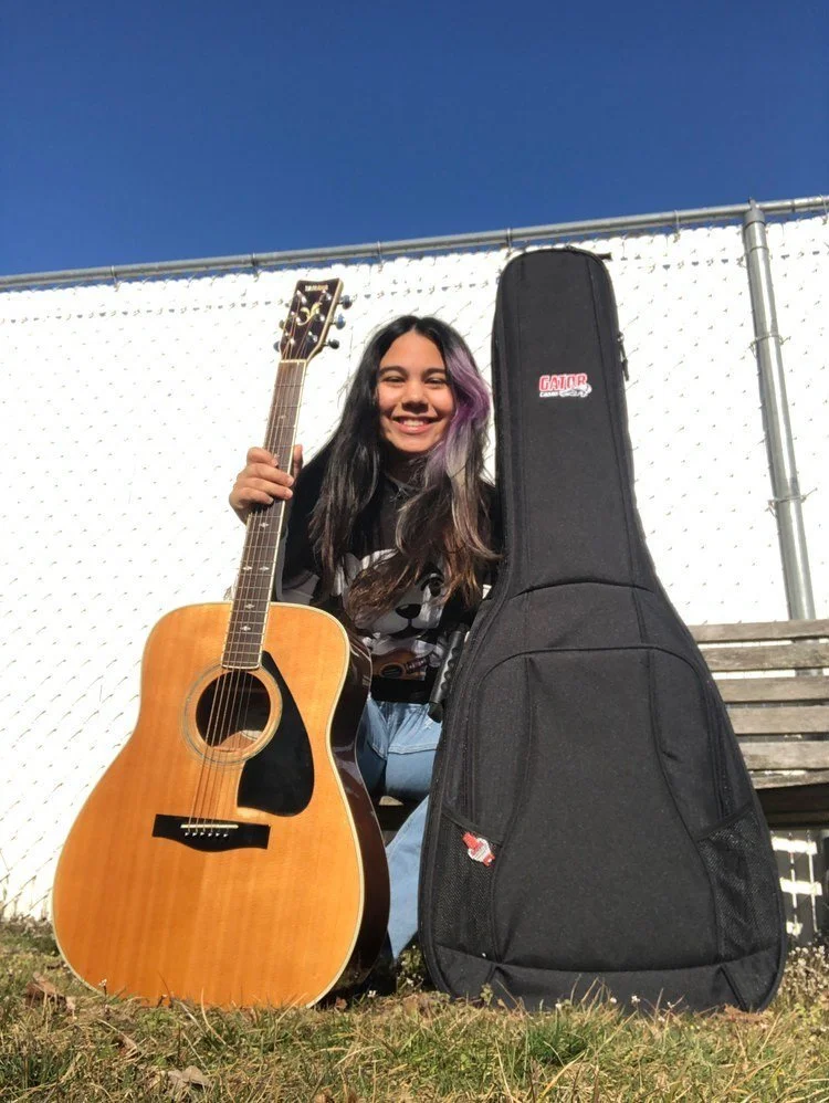 A young woman with long dark hair and purple streaks is smiling and holding an acoustic guitar. She is kneeling on the grass beside a black guitar case and a wooden bench, with a white fence and clear blue sky in the background.