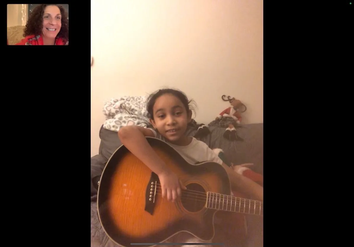 A young girl sitting on a couch holding an acoustic guitar, with a woman on a video call appearing in the top left corner. The girl is looking at the camera and has a slight smile. In the background, there is a stuffed elf on the sofa and a blanket.