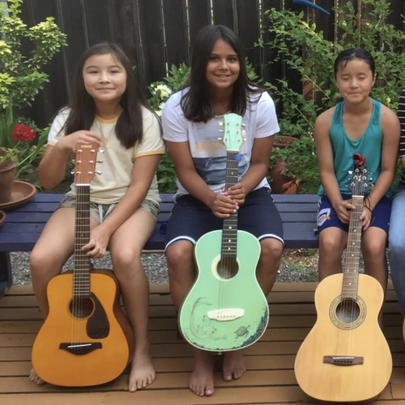 Three girls sitting on a bench in a backyard, each holding an acoustic guitar. The girl on the left has long dark hair, wearing a light yellow shirt and shorts. The girl in the middle has shoulder-length dark hair, wearing a white top with a striped 