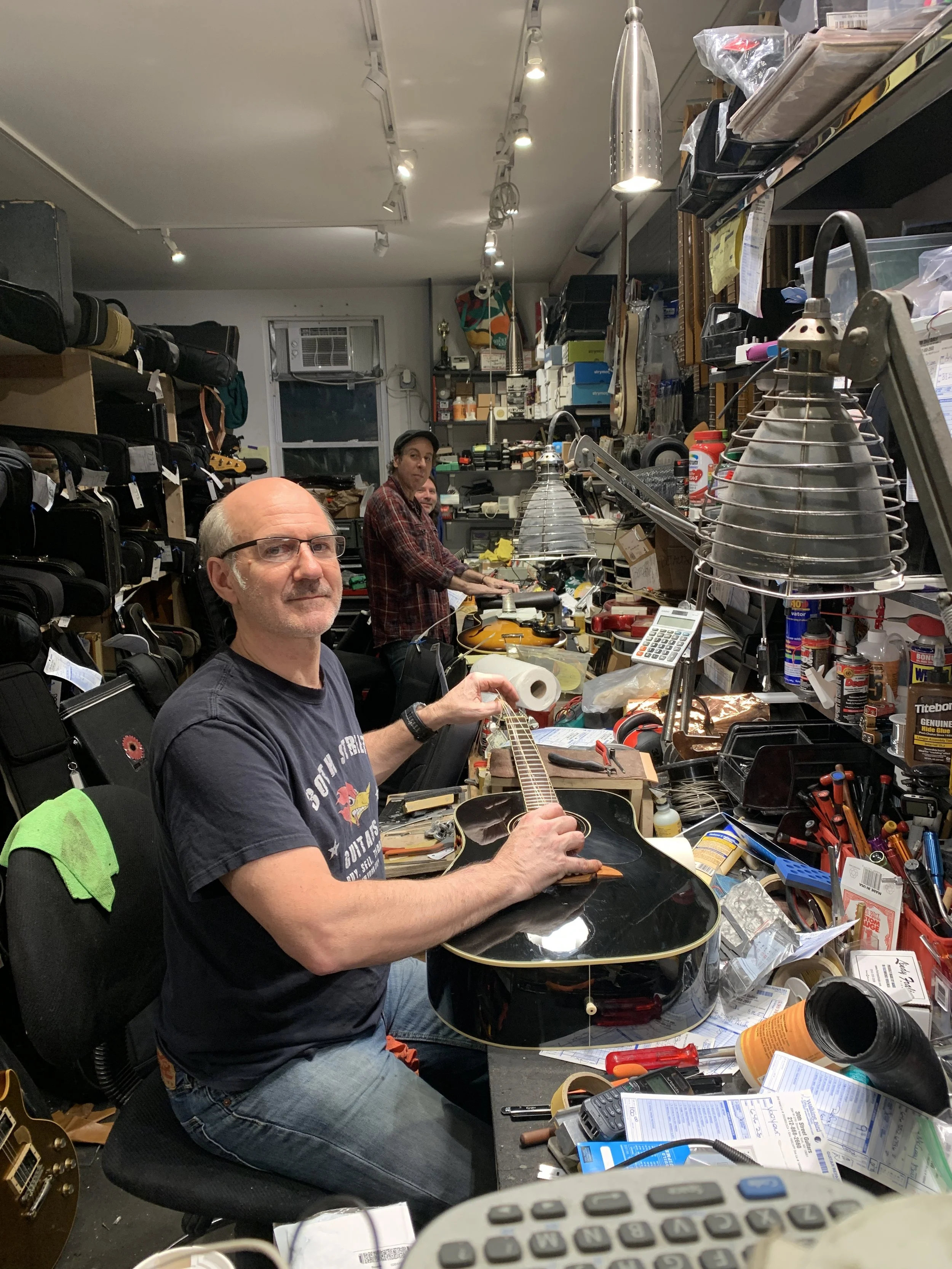 Man sitting at a cluttered workbench holding a black acoustic guitar, with two people in the background working amid tools and equipment in a busy workshop.