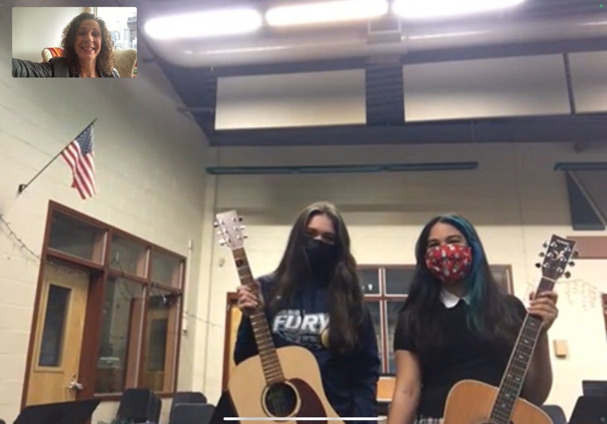 Two women wearing masks holding guitars inside a room with an American flag on the wall and chairs, smiling at the camera during a virtual meeting.