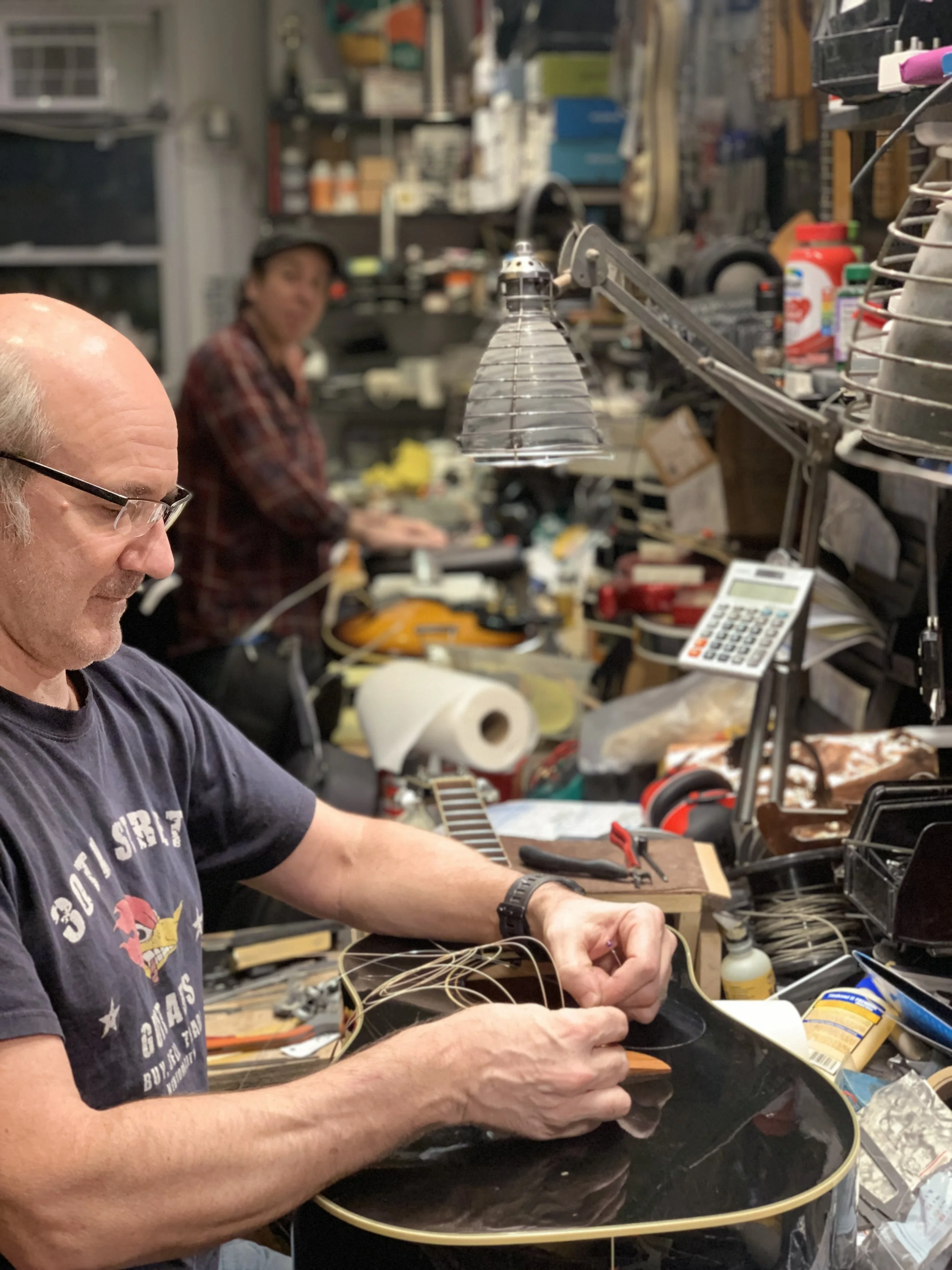 A man working on a guitar in a cluttered workshop, with a woman in the background, surrounded by various tools, supplies, and equipment.