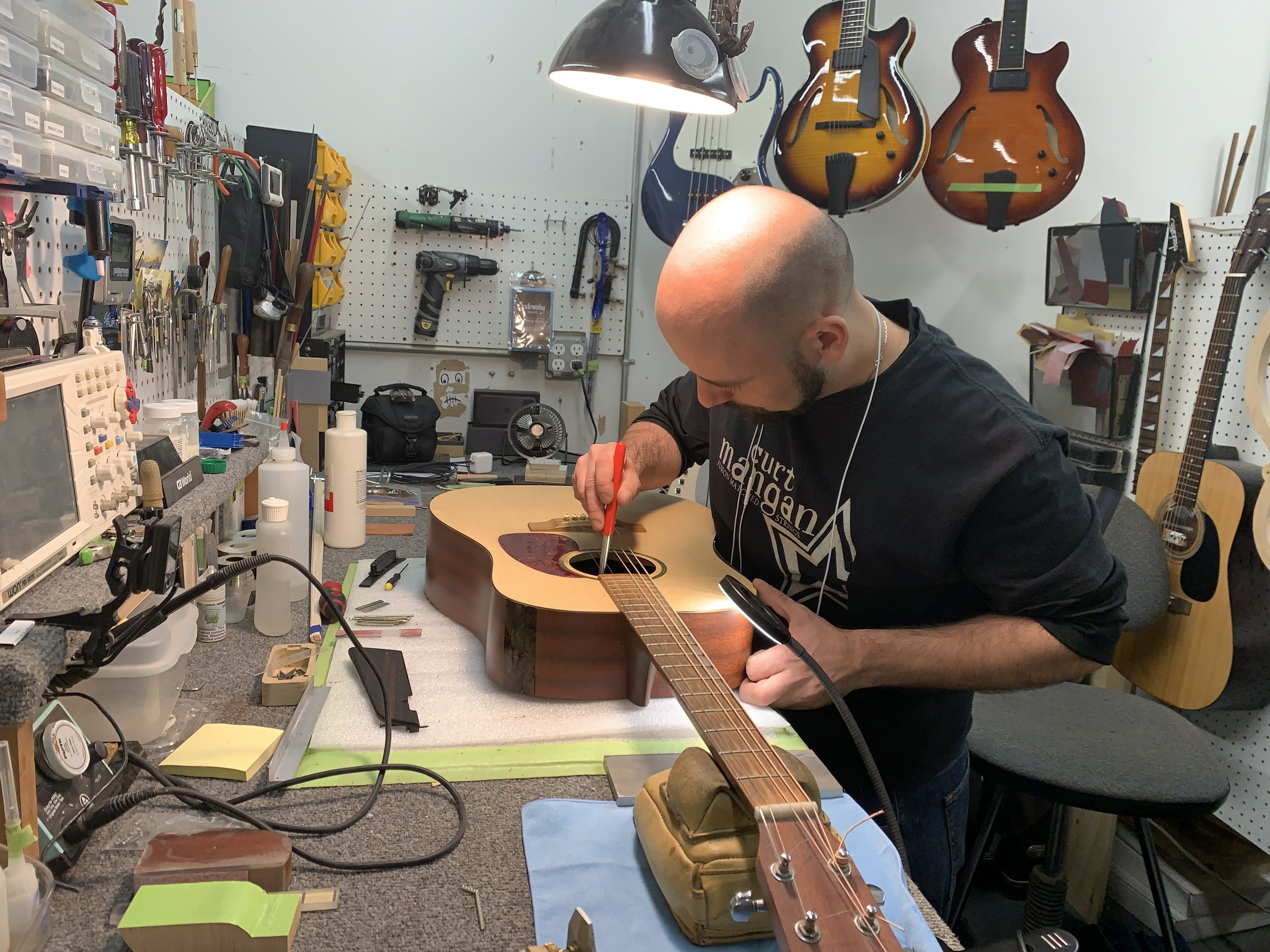 A person working on an acoustic guitar in a workshop, surrounded by tools and guitar parts, with several guitars hanging on the wall behind him.