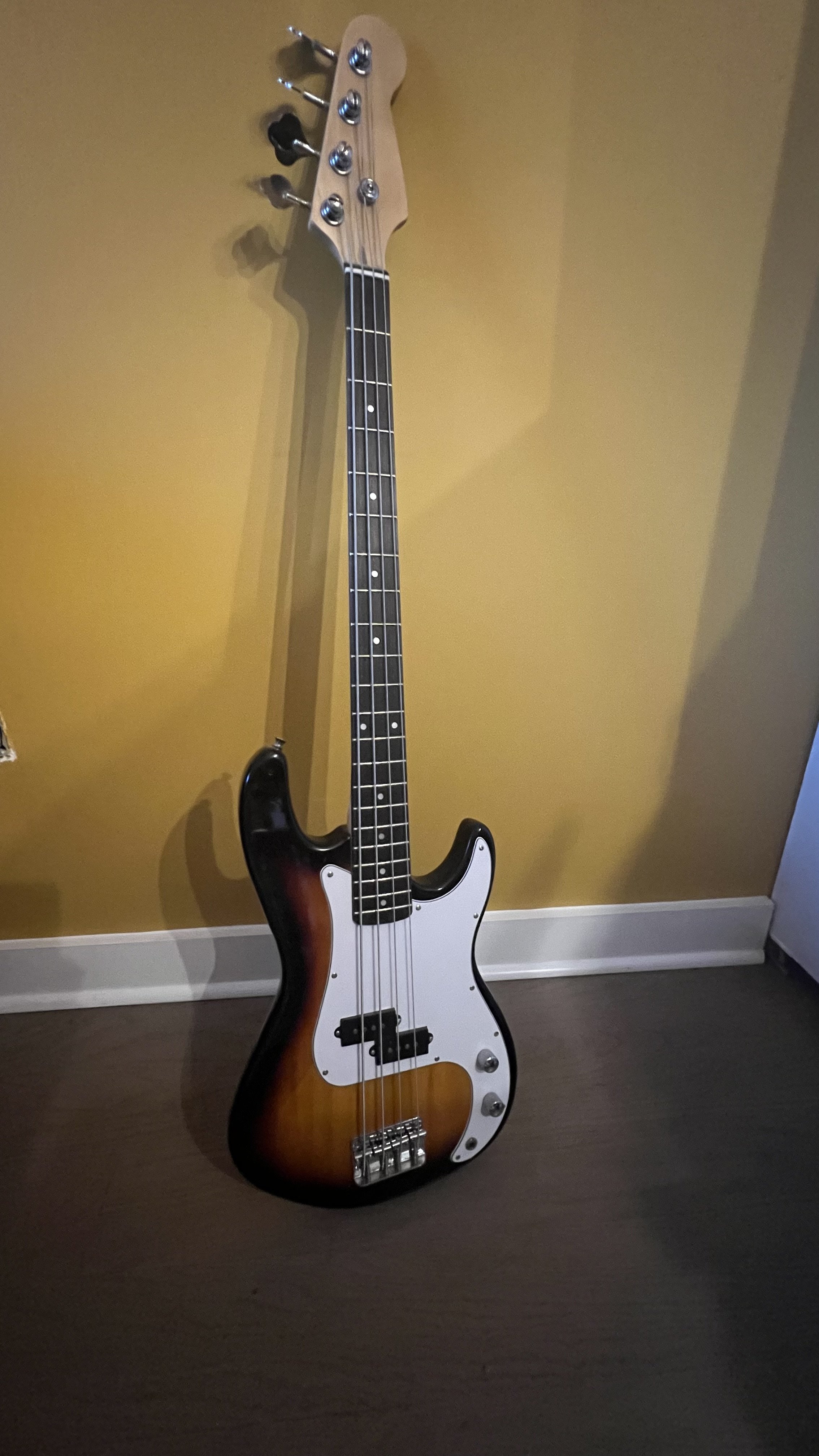 An electric bass guitar with a sunburst finish, white pickguard, and four strings, leaning against a yellow wall on a brown floor.