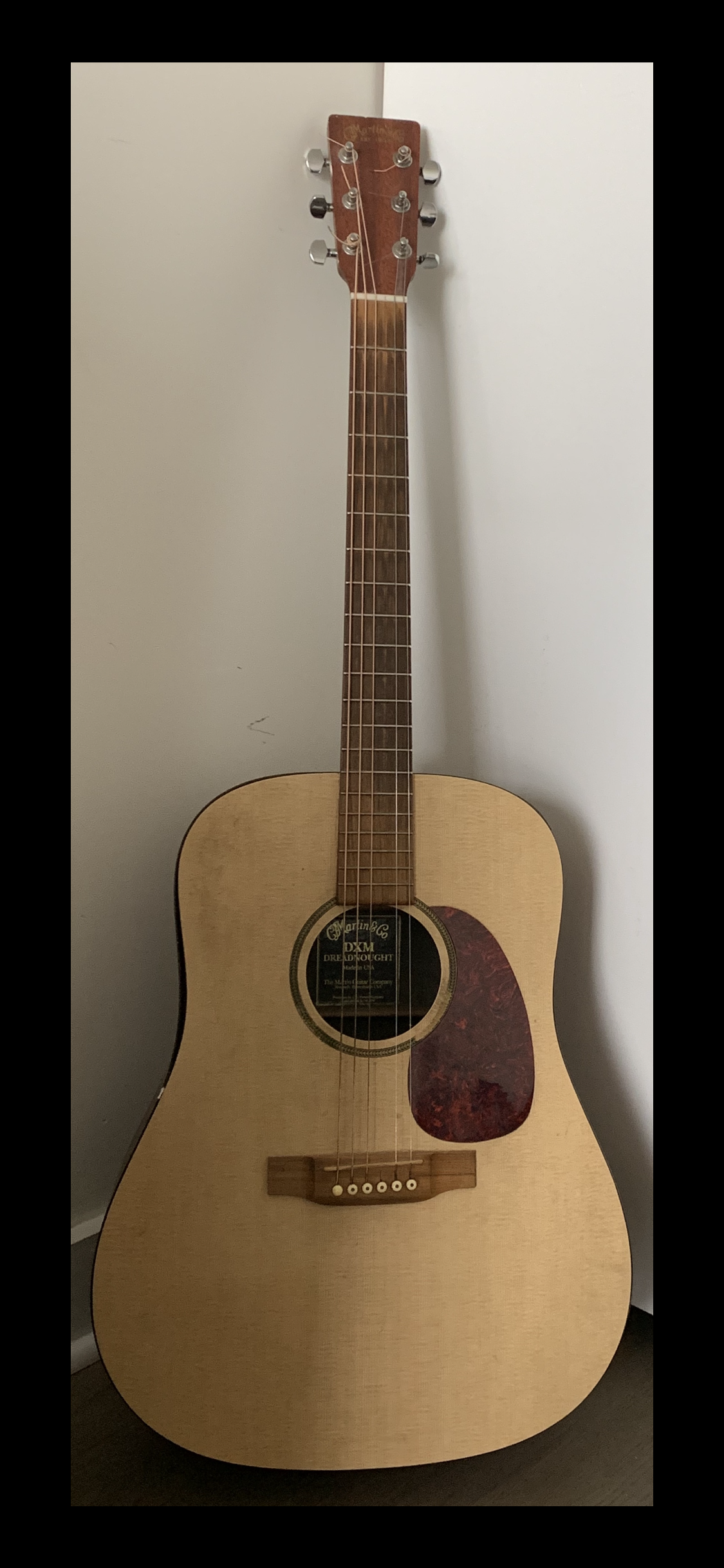 An acoustic guitar with a light wood body, dark wood neck, and chrome tuning pegs, positioned against a plain wall.