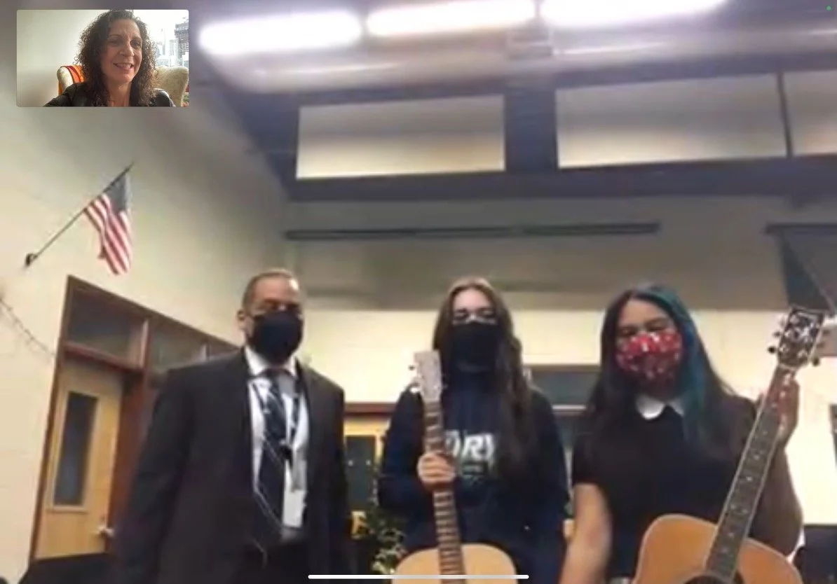 Three people standing in a room, two women and one man, all wearing masks and holding guitars, with an American flag on the wall to the left.