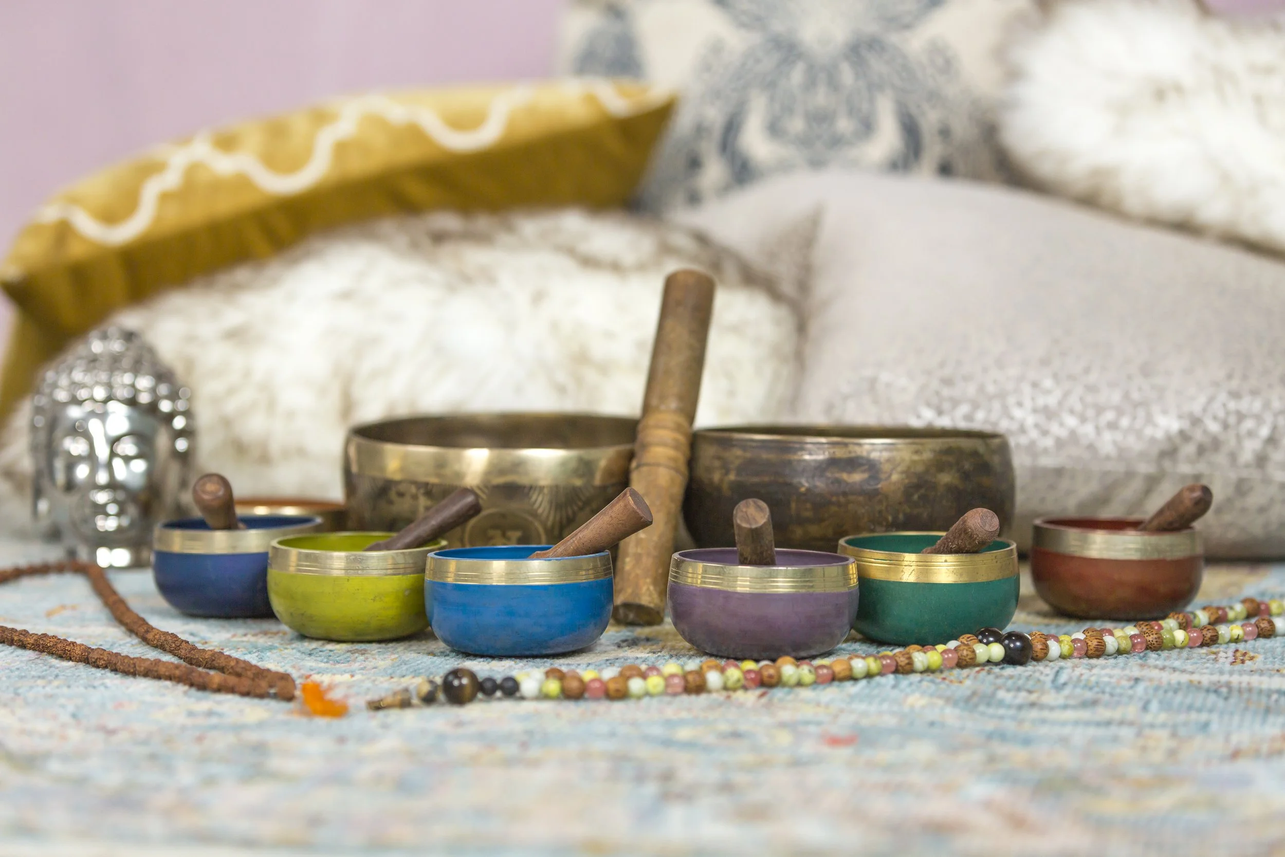 Colorful Tibetan singing bowls and mallets arranged on a patterned cloth, with beads and pillows in the background.