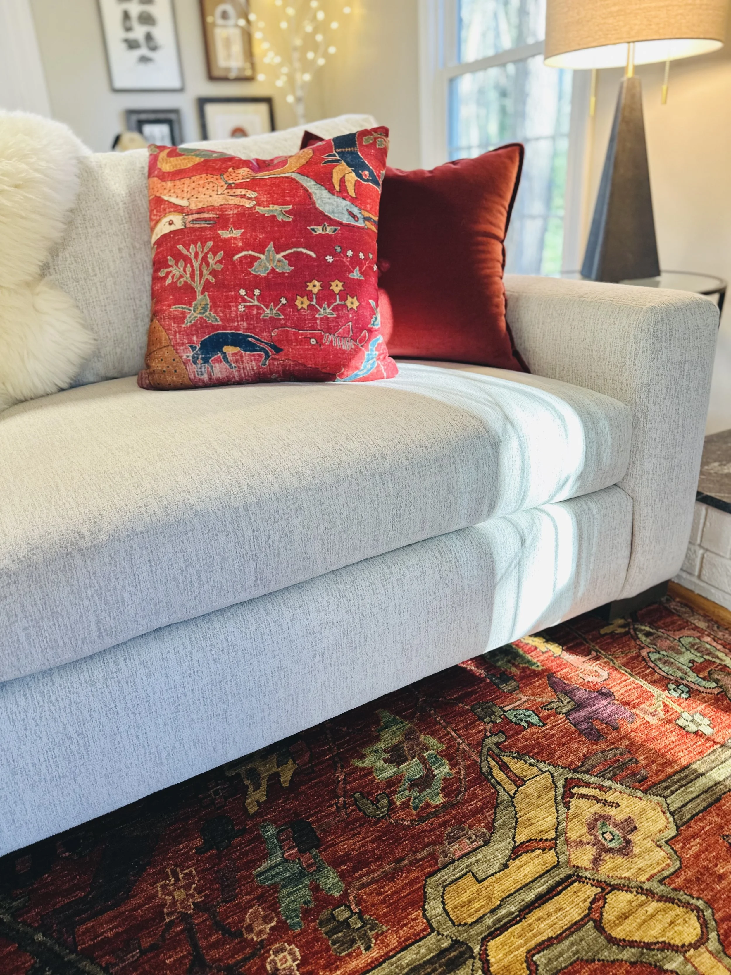 A living room with a light-colored sofa adorned with three pillows: one with a colorful, intricate pattern, and two in solid red. There's a nearby table with a standing lamp, and framed artwork decorates the wall. A patterned rug covers the floor.