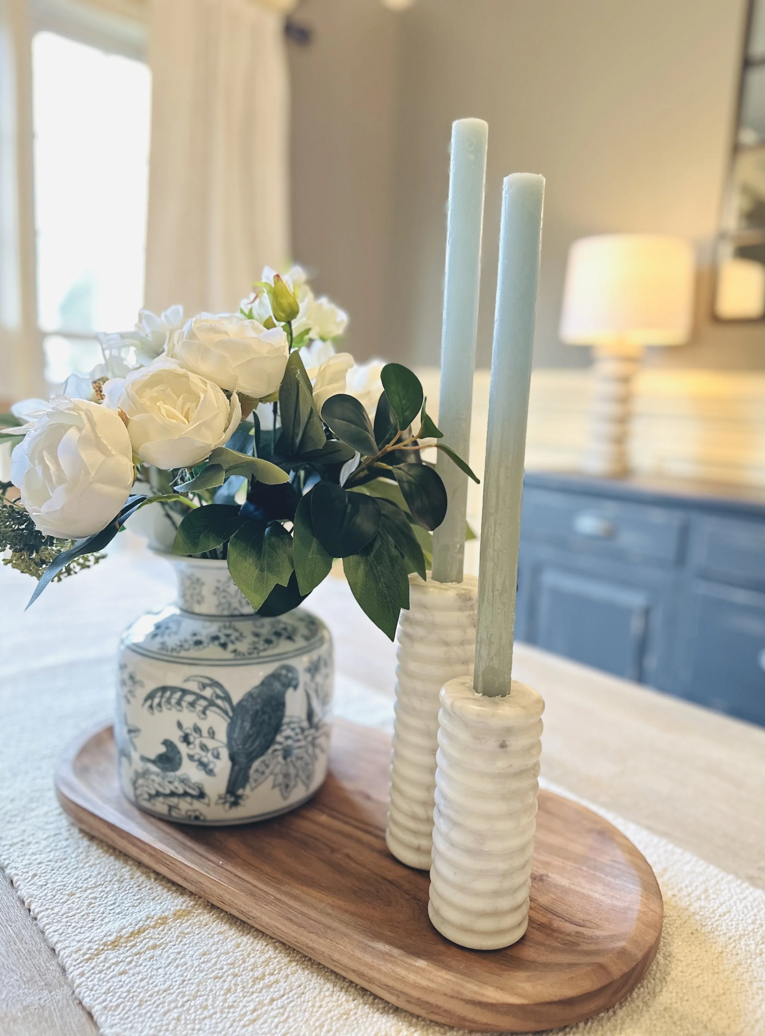 Decorative arrangement with a potted white flower and two light blue candles in ceramic holders on a wooden tray, placed on a table.