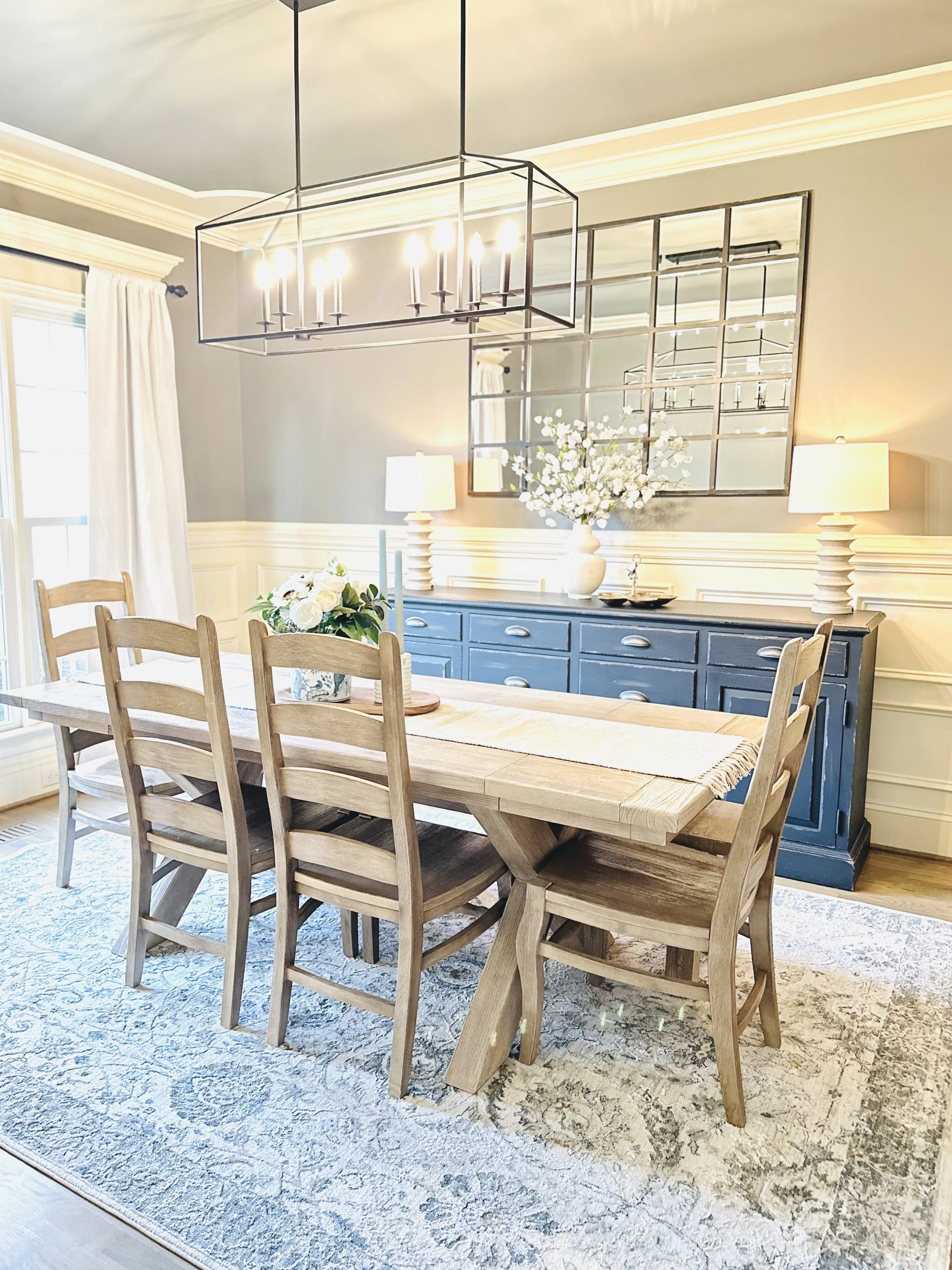 A dining room with a wooden table, six chairs, a blue sideboard, white lamps, and floral arrangements. A modern chandelier hangs above the table, and a mirror is on the wall behind the sideboard.