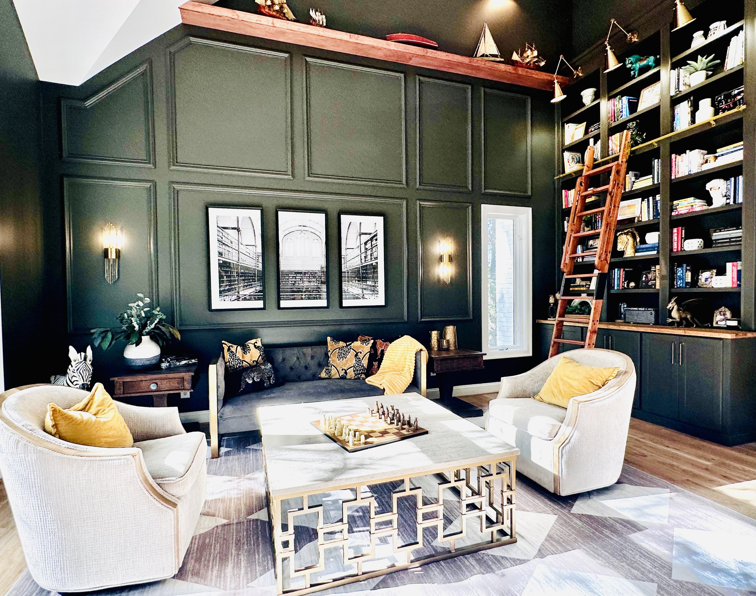 Living room with dark green paneled walls, two white armchairs with yellow pillows, a gray sofa with patterned pillows, a white coffee table with a chess game, a large built-in bookshelf with a wooden ladder, a window, and modern wall sconces.