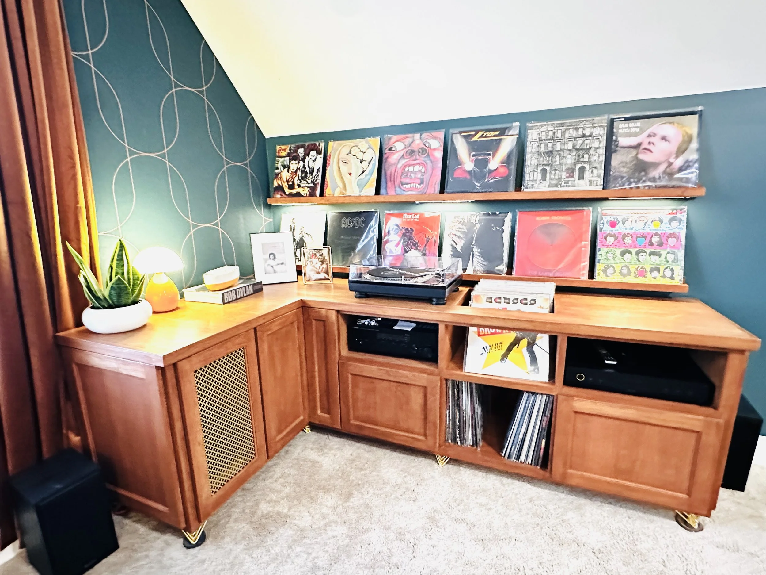 Corner wooden cabinet with vinyl records on shelves, turntable on top, small lamp, potted plant, and framed photos, against a wall with a blue patterned wallpaper and a yellow curtain.