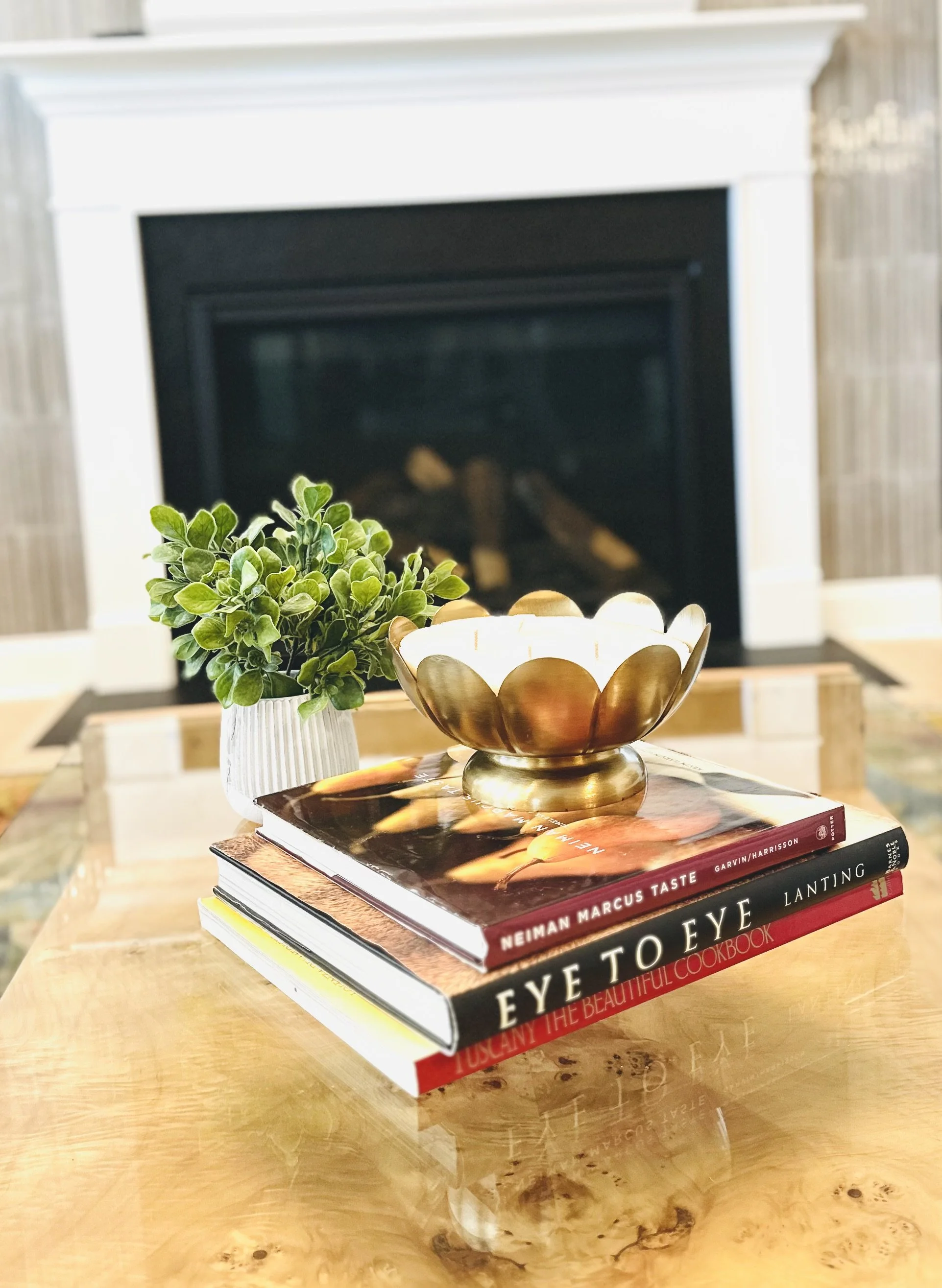 A wooden table with a small potted green plant, a decorative gold and white bowl, and three books titled "Neiman Marcus Taste," "Eye to Eye," and "Tuscan: The Beautiful Cookbook" in front of a fireplace.