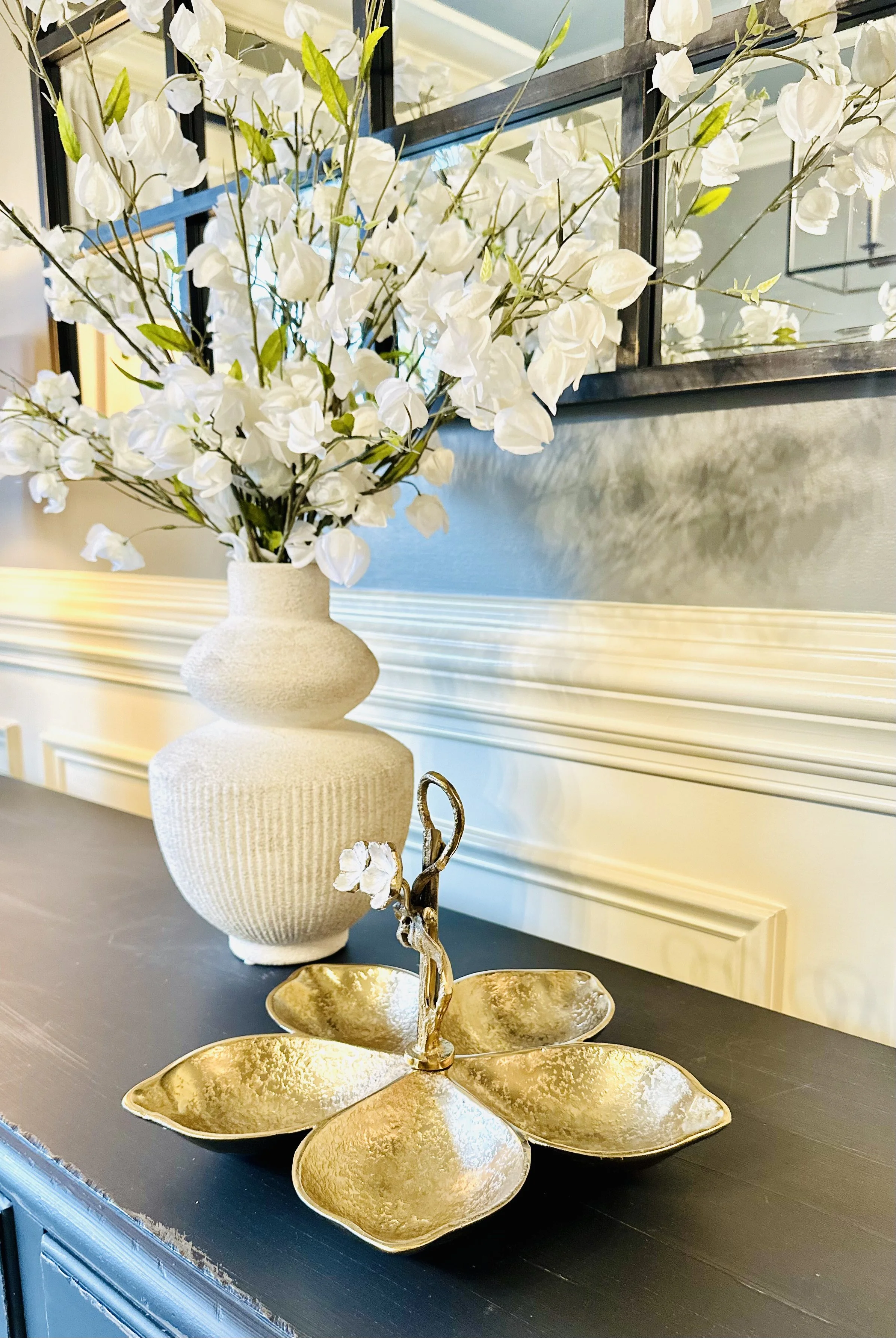 A decorative arrangement featuring a large white textured ceramic vase with white flowers and green leaves, and a gold-colored royalty-inspired decorative piece resembling a lotus flower with a human figure on top, on a dark wooden table in a well-decorated interior.