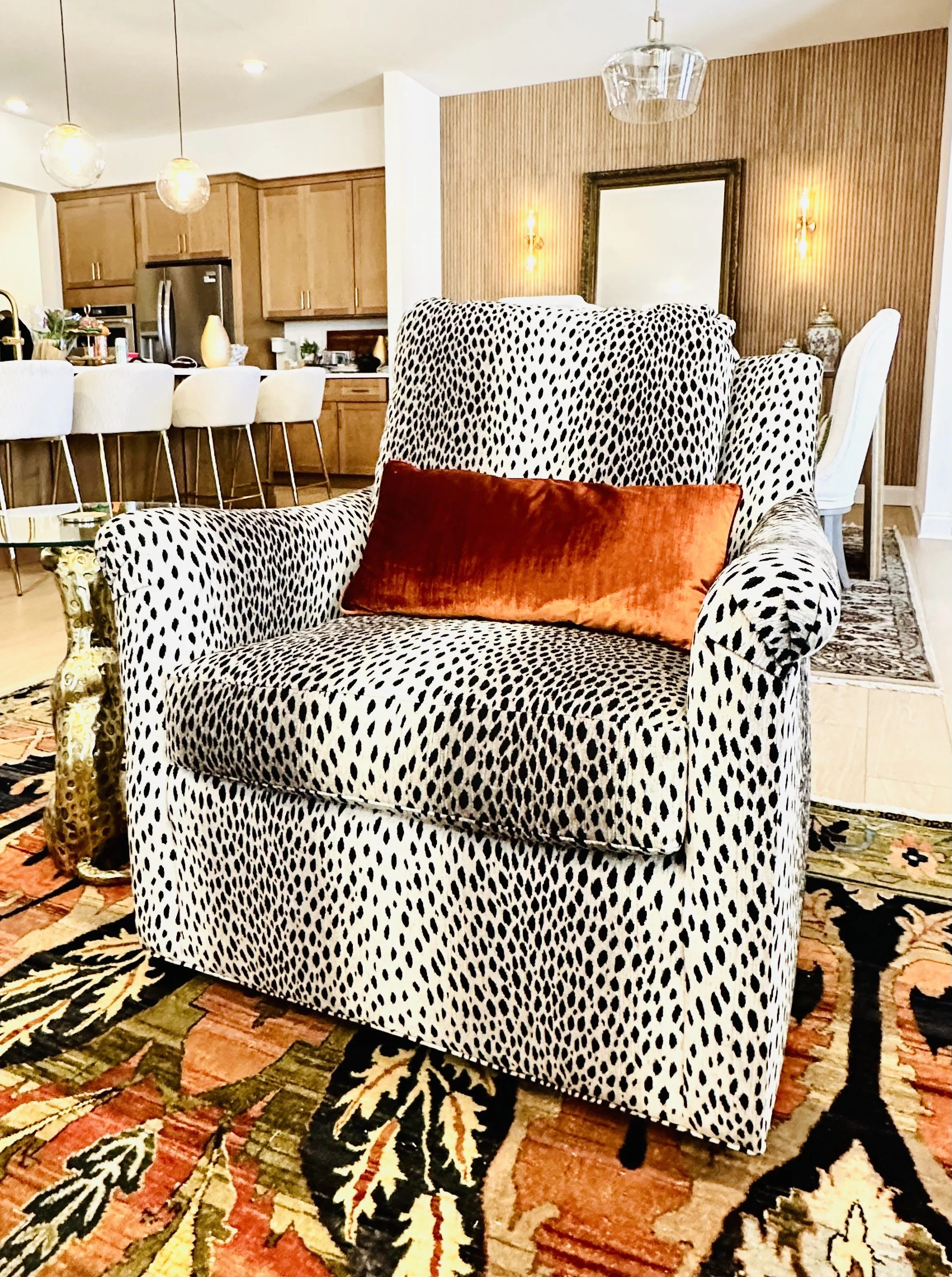 Living room with a black and white patterned armchair, orange velvet pillow, patterned area rug, kitchen with wooden cabinets and white chairs in the background, decorative items, and wall-mounted lights.