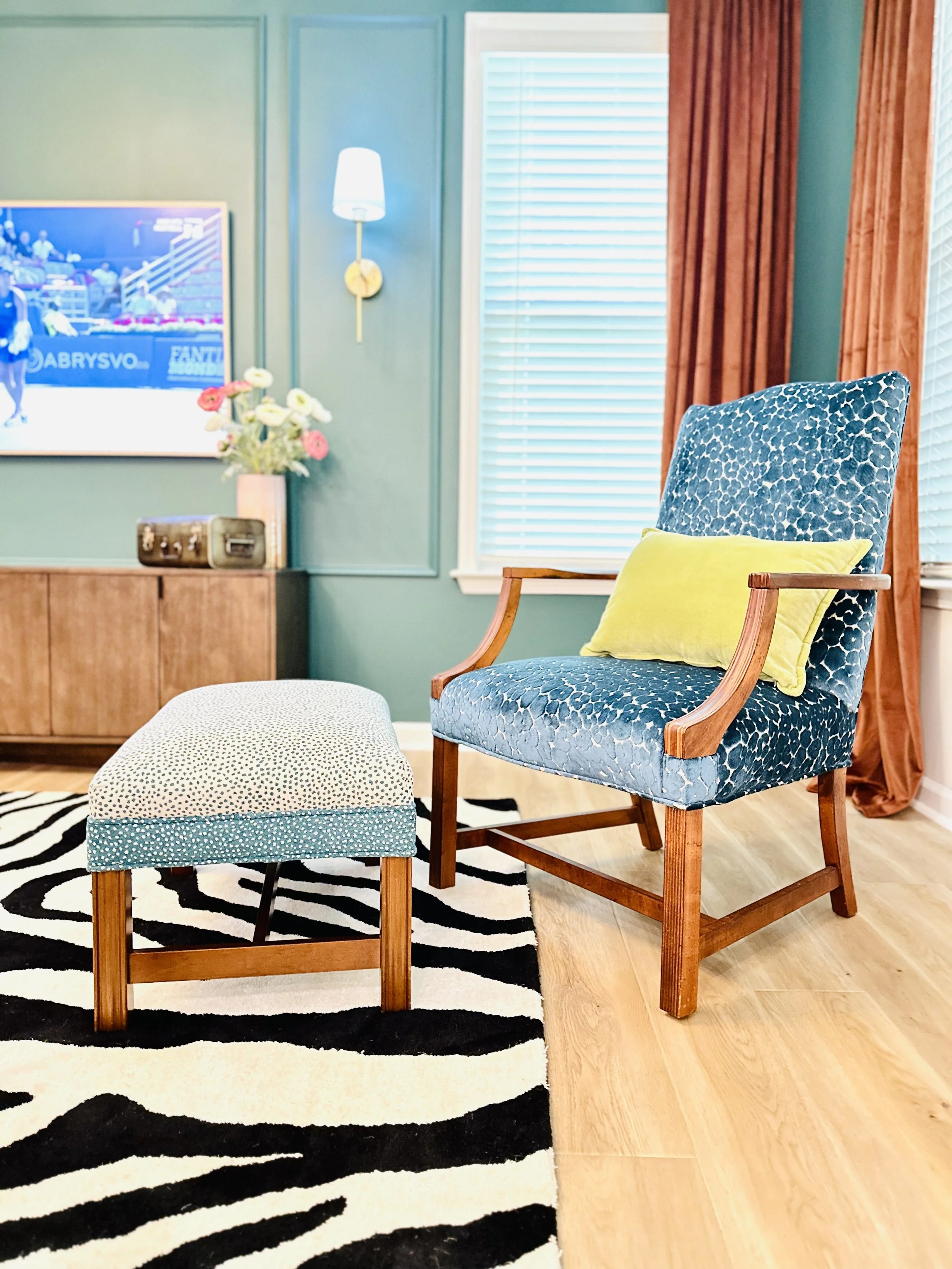 A living room with a blue patterned armchair, yellow pillow, matching ottoman, zebra-print rug, and teal wall with a wall-mounted light and a flat-screen TV.
