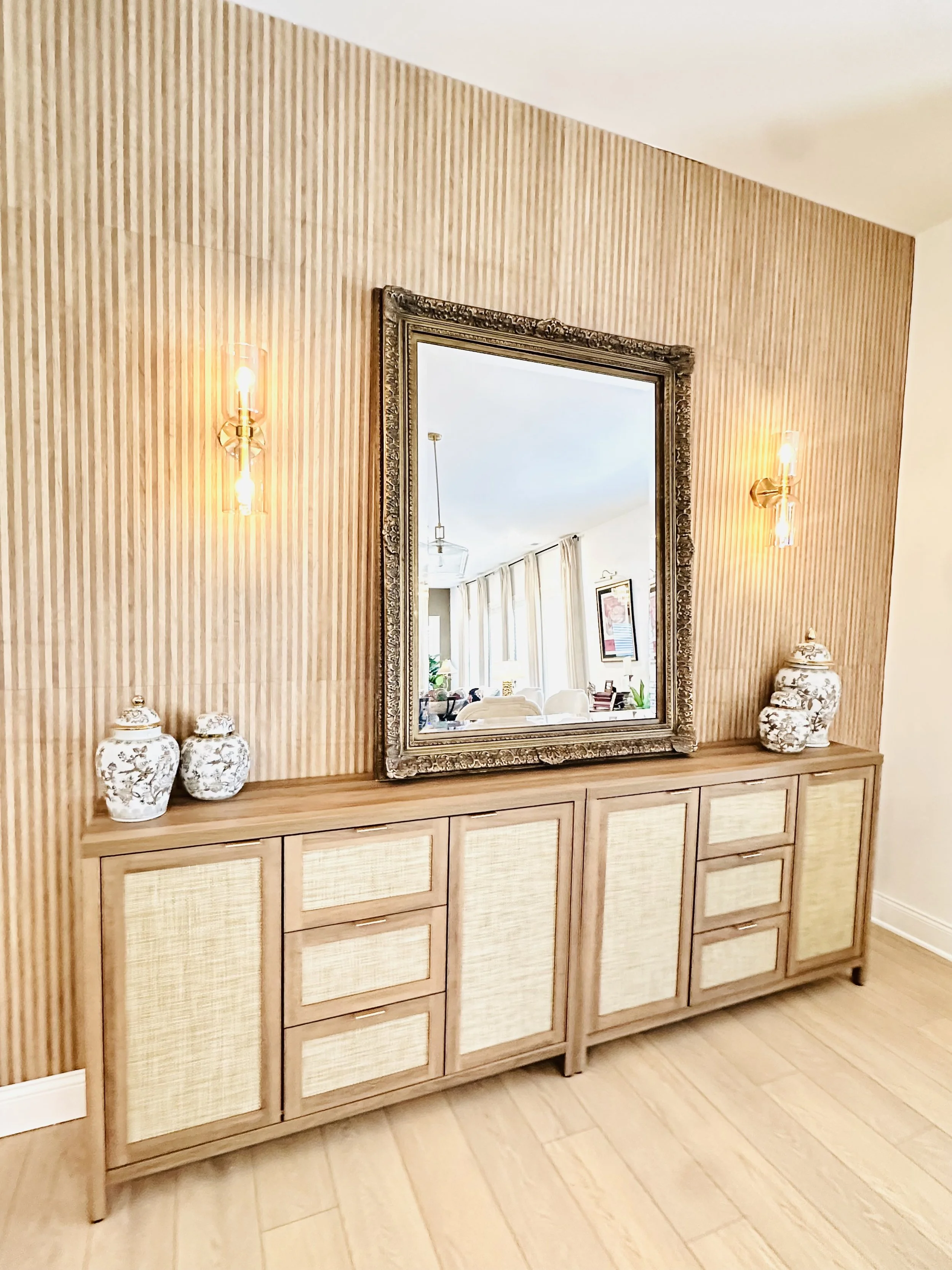 A wooden sideboard with six drawers and two cabinets, topped with two decorative porcelain vases, stands against a textured beige wall with vertical lines. A large ornate mirror is centered above the sideboard, and two brass wall sconces with exposed bulbs are mounted on either side of the mirror, illuminating the space.