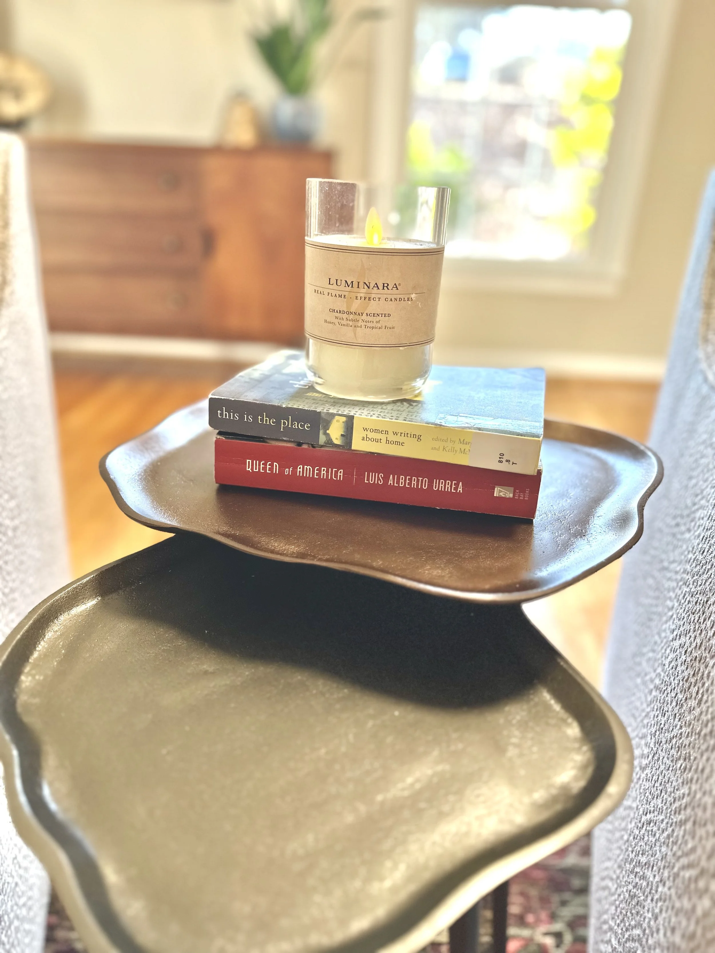 A candle labeled Luminar is placed on top of two stacked books on a small table, with a background of a sunlit room. The books are titled "this is the place" and "Queen of America".