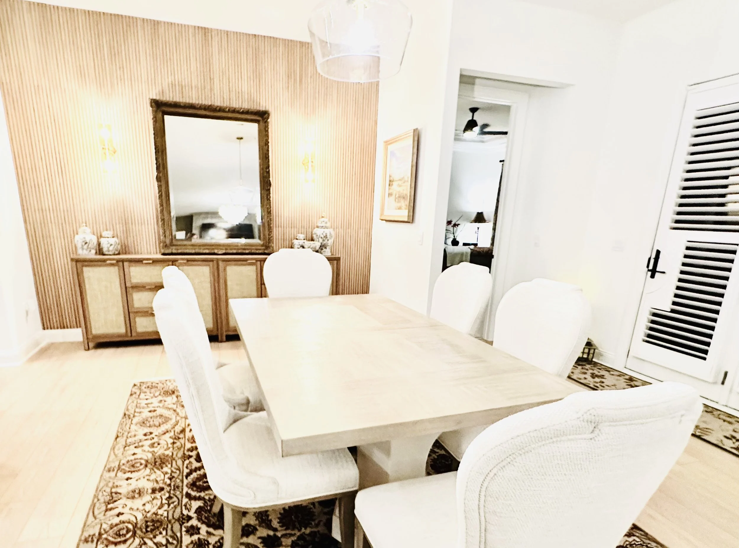 Dining room with a large rectangular table surrounded by six white upholstered chairs, a wooden sideboard with decorative vases, a large mirror on a wooden paneled wall, and white walls with a doorway leading to another room.