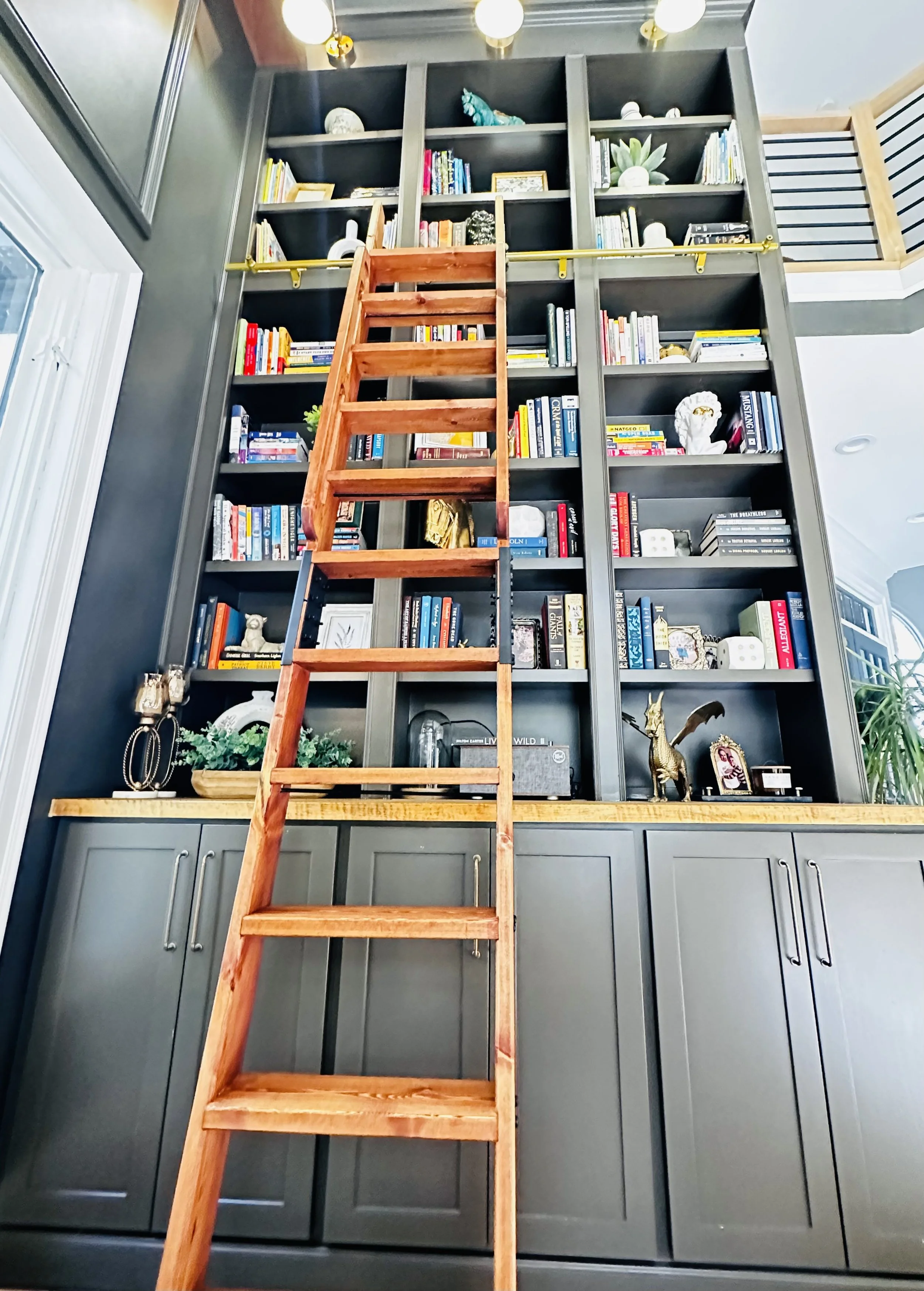 Tall dark gray bookshelf filled with books, decorative objects, and plants, with a small wooden ladder leaning against it.