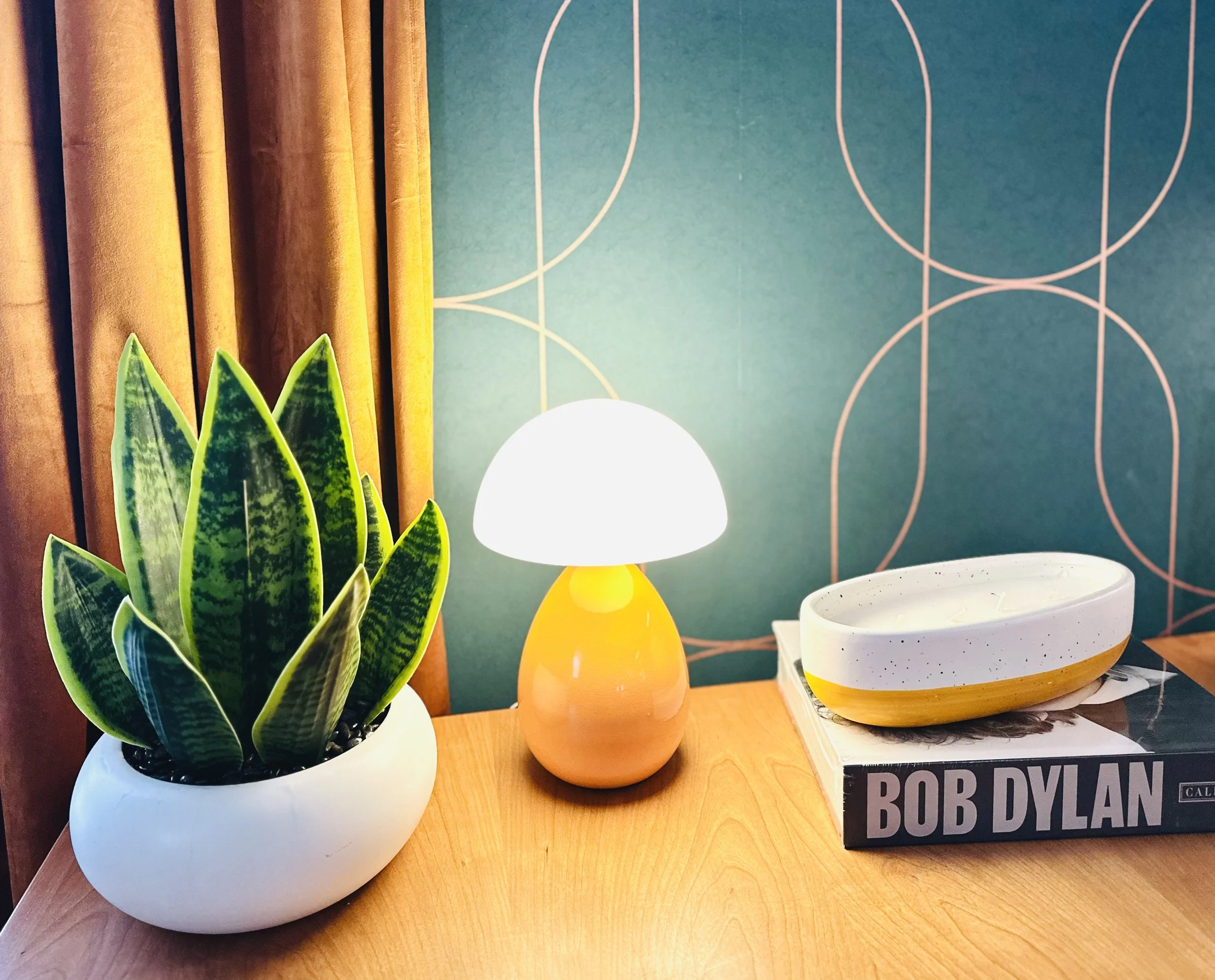 Decorative table with a potted snake plant, a small table lamp, a ceramic bowl, and a book titled 'Bob Dylan,' set against a blue wall with a geometric pattern.