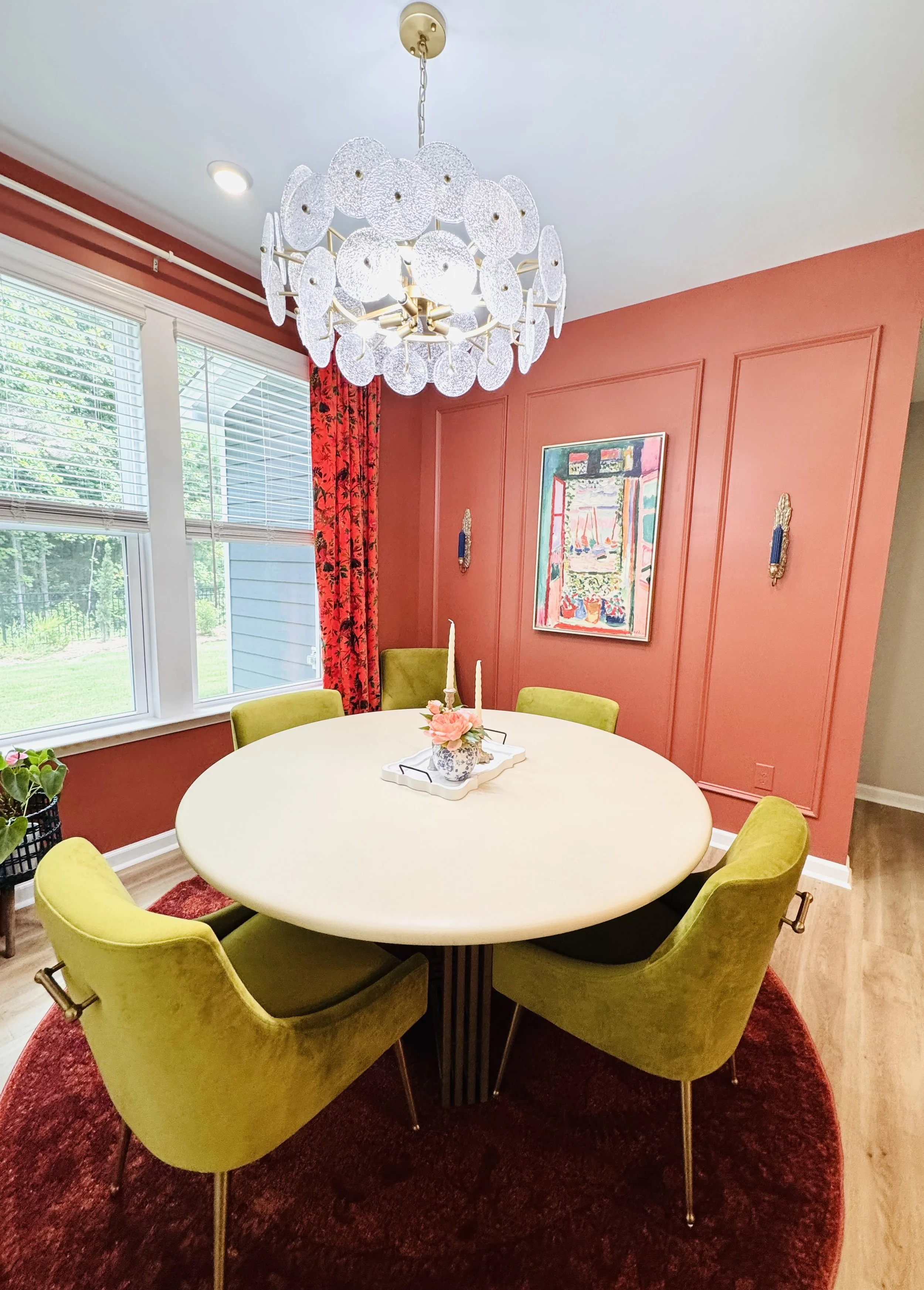 A dining room with a round white table surrounded by six green velvet chairs. The room features a red wall with a colorful painting, window with blinds, red curtains, a red circular rug, and a modern white chandelier hanging from the ceiling.
