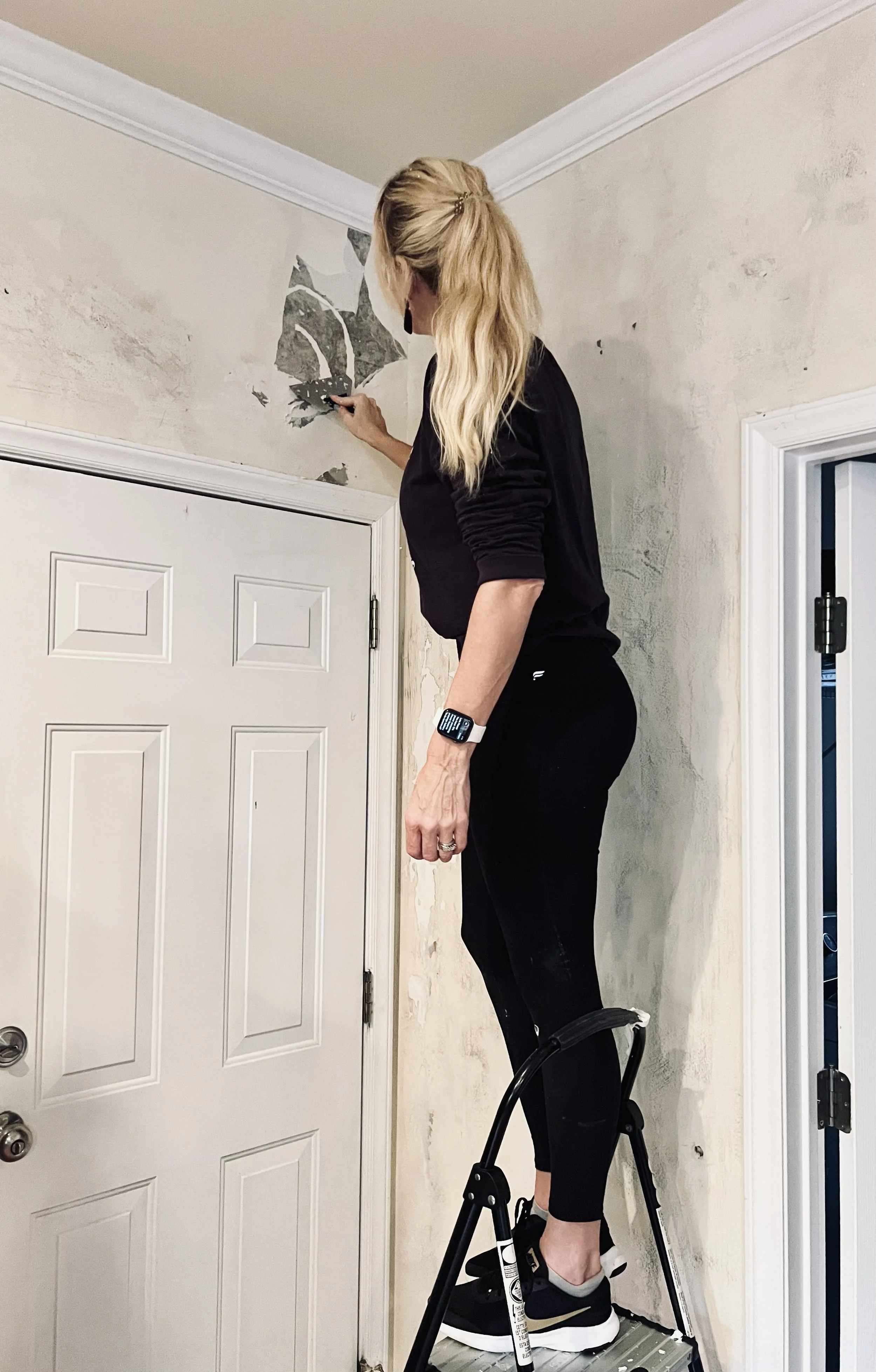 A woman standing on a step stool removing wallpaper from a wall in a room with white trim and a closed door.