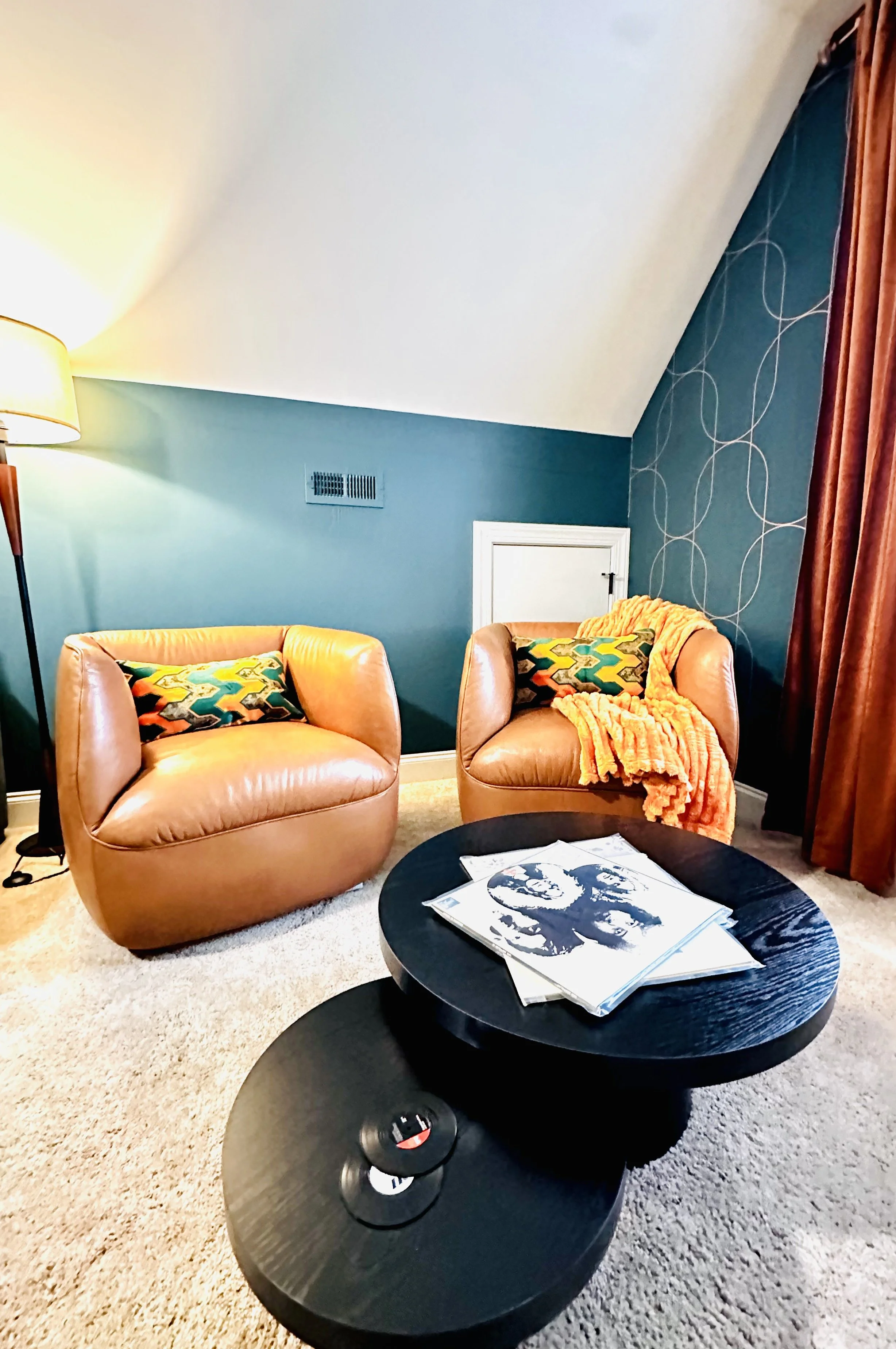 A cozy living room corner with two tan leather chairs, one with a colorful pillow, and an orange blanket. A black coffee table with vinyl records on top. A tall floor lamp, teal wall with pattern, and a small white door in the background.