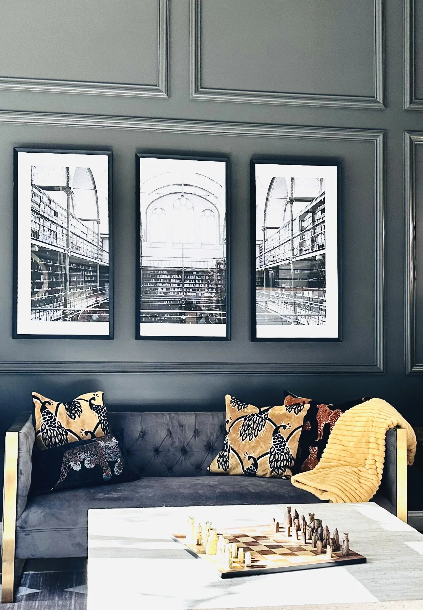 A gray sofa with three decorative pillows and a yellow throw blanket, a chessboard with chess pieces on a table in front, and three framed black and white photos of a library interior on a gray paneled wall.