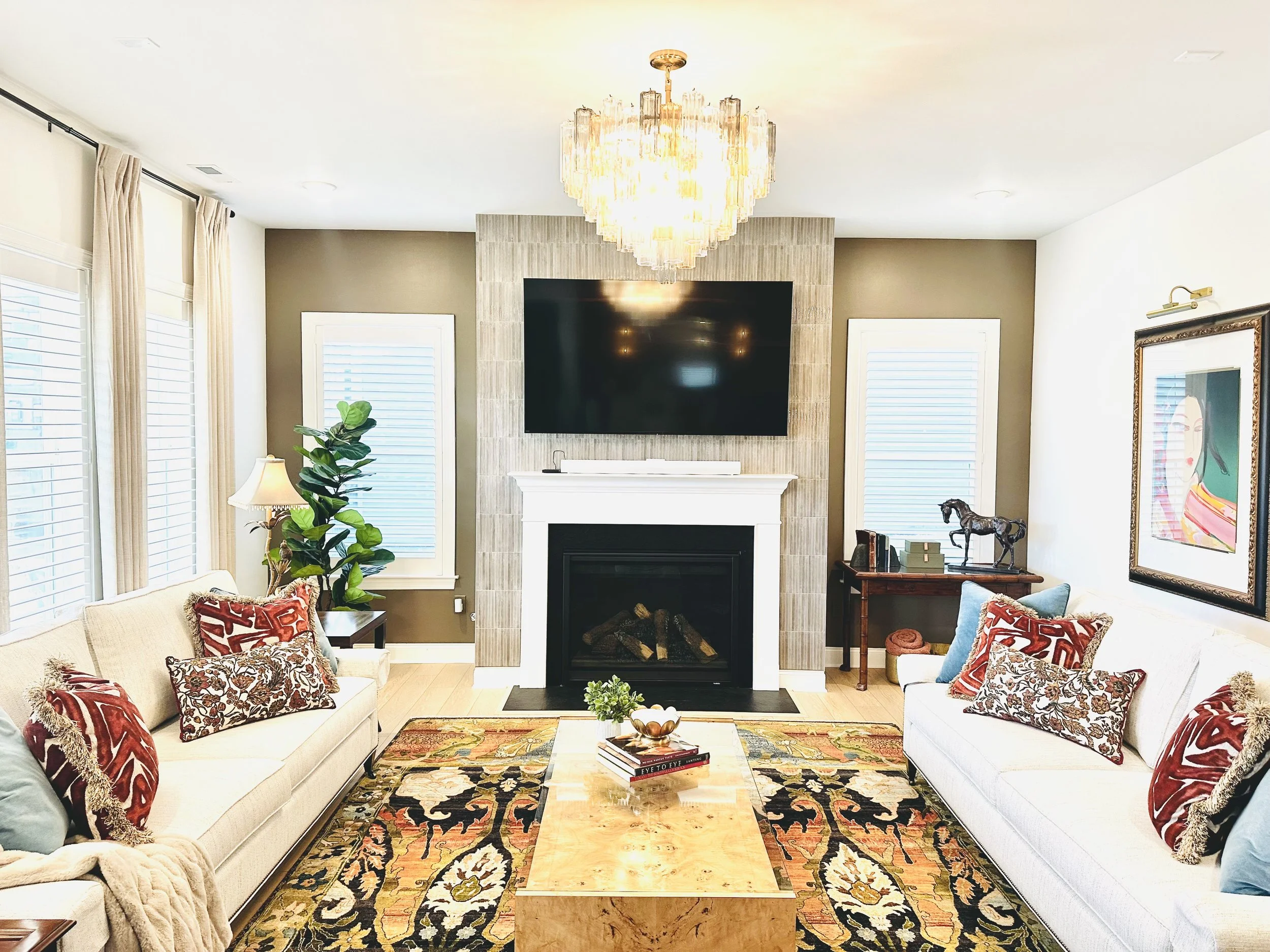 Living room with two white sofas, colorful patterned pillows, wooden coffee table, fireplace with a large wall-mounted TV, chandelier, windows with curtains, potted plant, framed artwork, and decorative items.