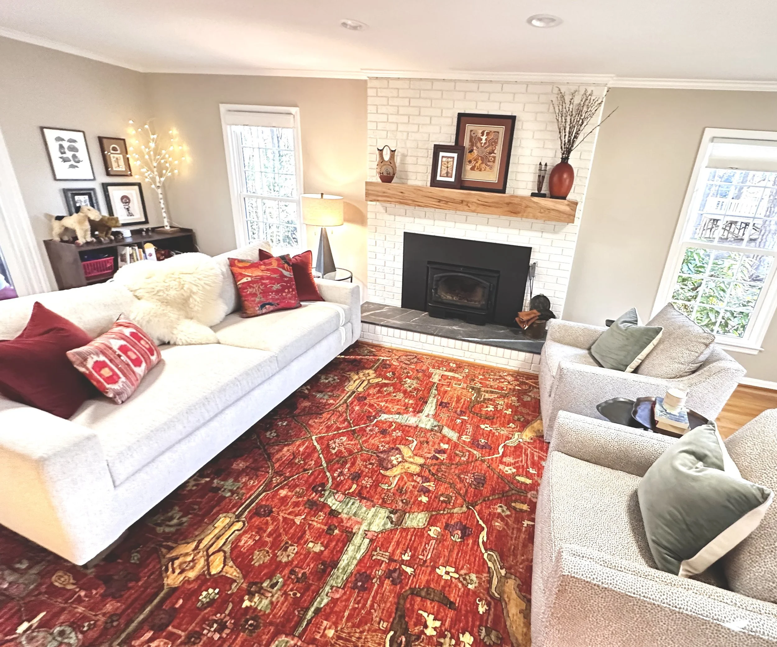 Living room with a white sofa, beige armchairs, a fireplace with a brick surround and a wooden mantel, a patterned red rug, and large windows.