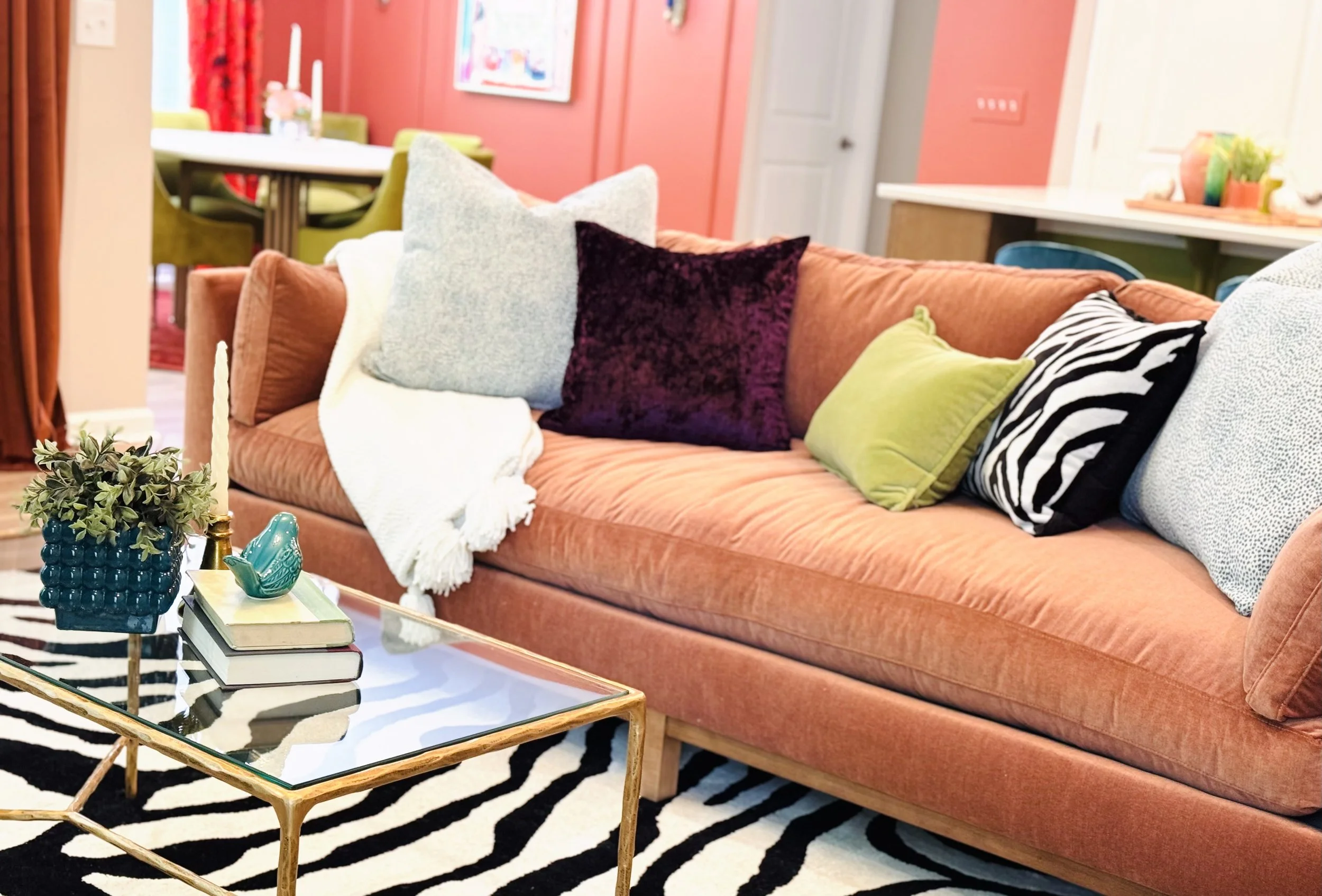 Living room with a blush pink sofa adorned with various decorative pillows, a glass coffee table with books and a plant, zebra print rug, and a colorful dining area in the background.