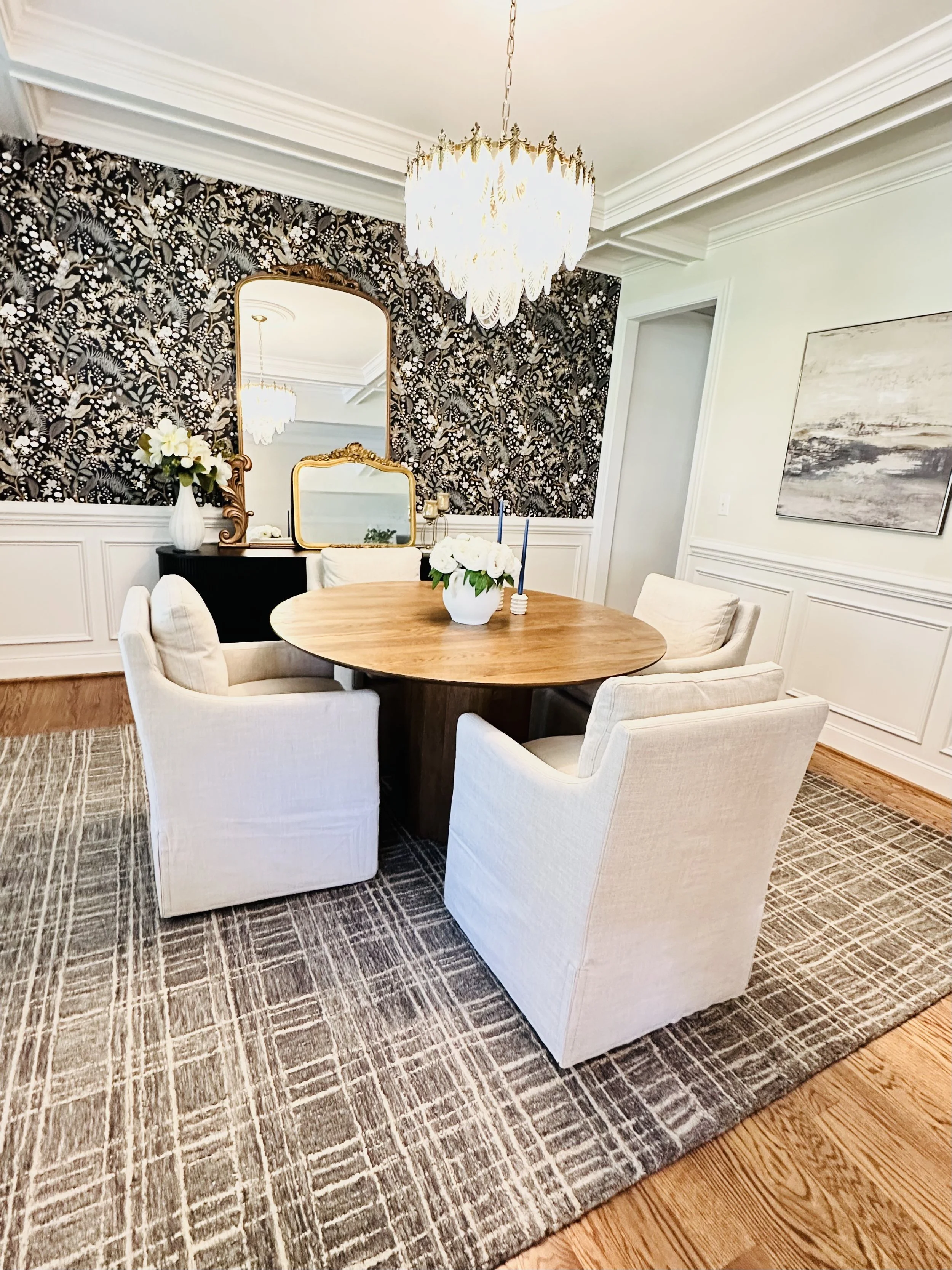 A dining room with four cream upholstered chairs around a round wooden table. The table has a white vase with white flowers and two blue candles. The room features black and white floral wallpaper on one wall, a large ornate mirror, and a chandelier hanging from the ceiling. There's also a black sideboard with a white vase of flowers and a gold-framed mirror above it, and white wainscoting on the walls.