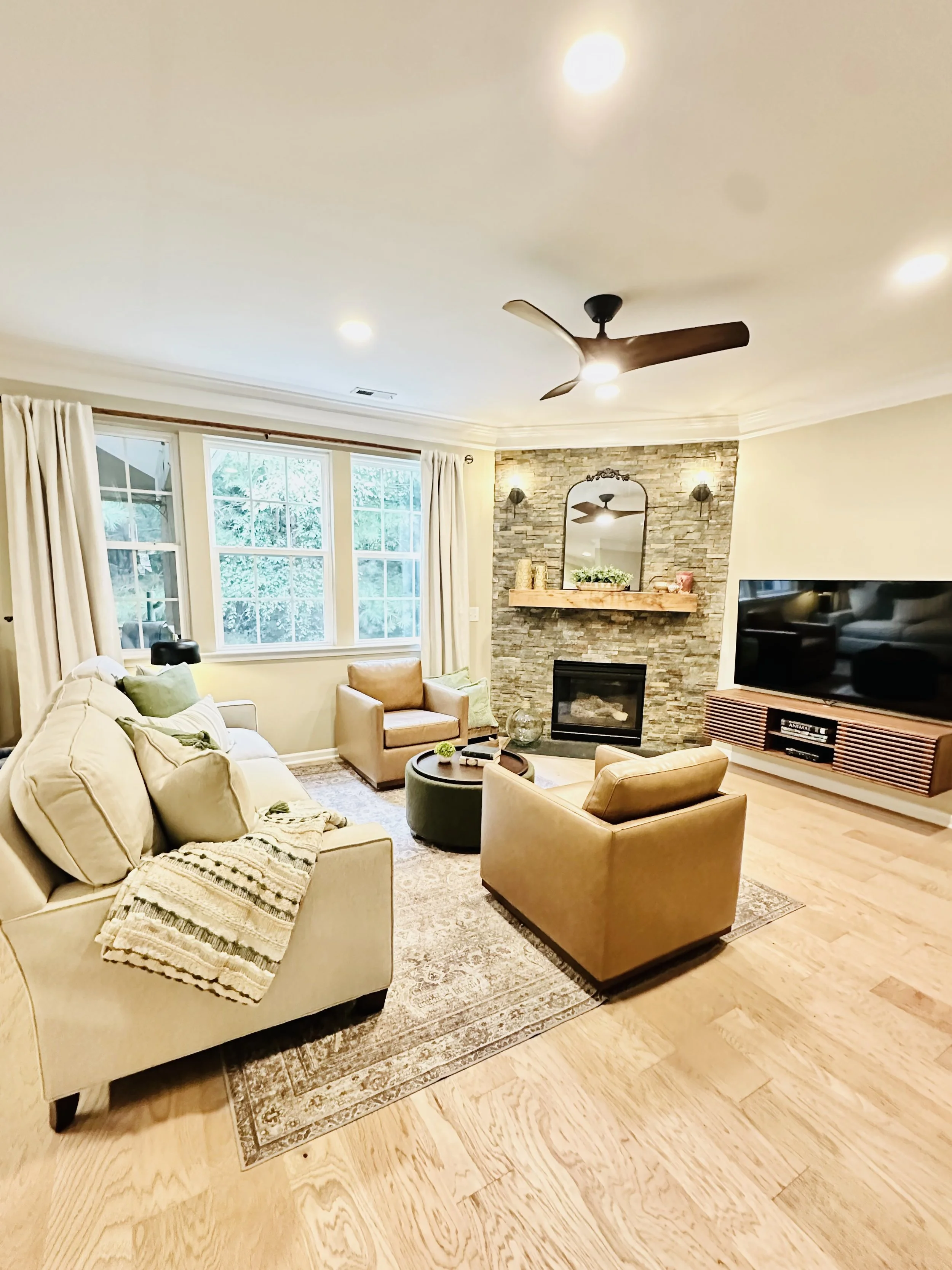 Living room with beige sofa, tan armchairs, a green ottoman, a fireplace with stone surround, a large flat-screen TV, and large windows with white curtains.