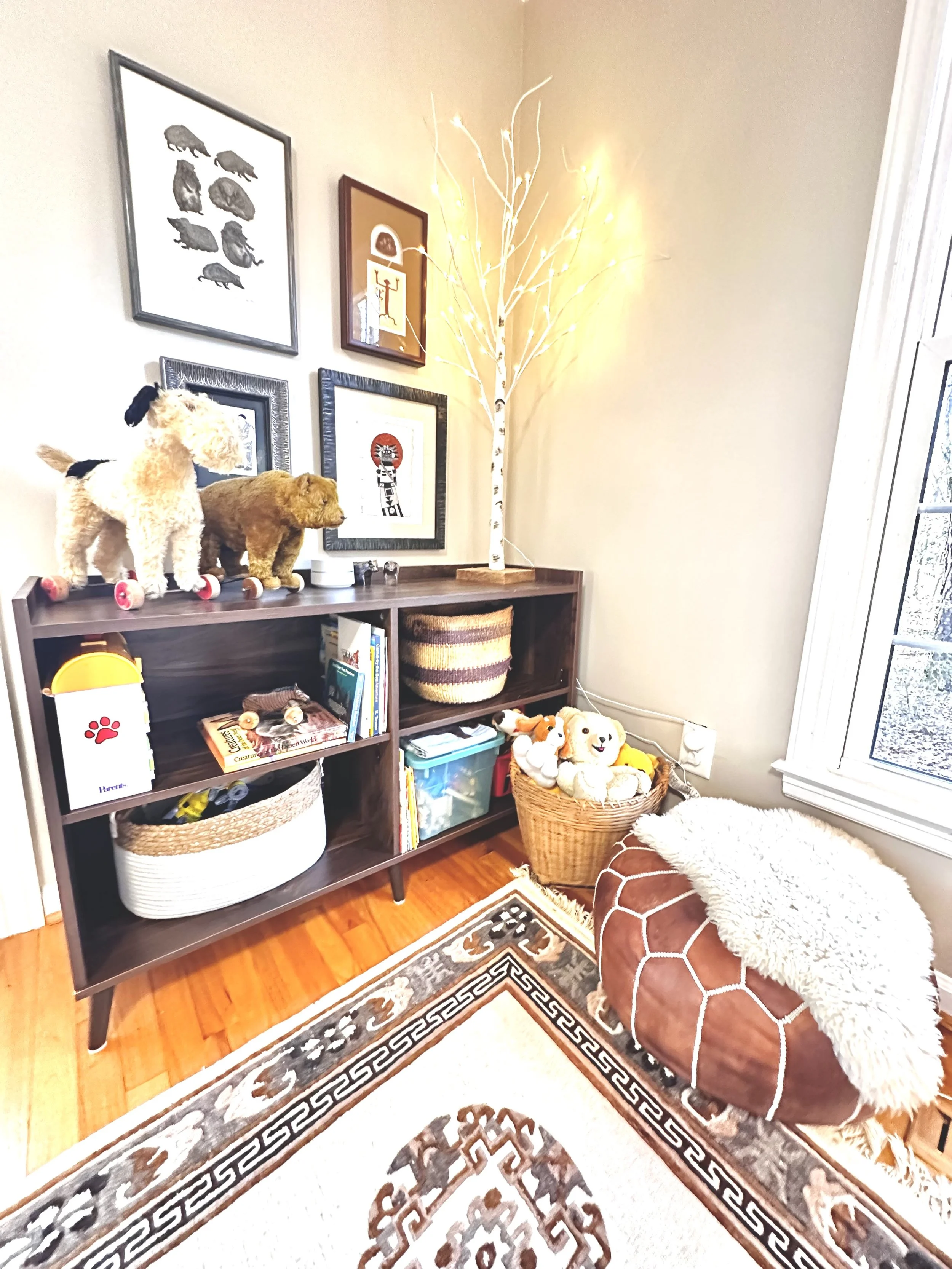 A cozy living room corner with a wooden bookshelf filled with books and baskets, plush dog toys, and a decorative basket of stuffed animals. A white illuminated artificial tree stands beside the shelf, casting a warm glow. The wall is decorated with framed artwork and photographs. A floor rug with a geometric pattern covers part of the wooden floor, and a furry pouf is next to a powdery pink blanket.