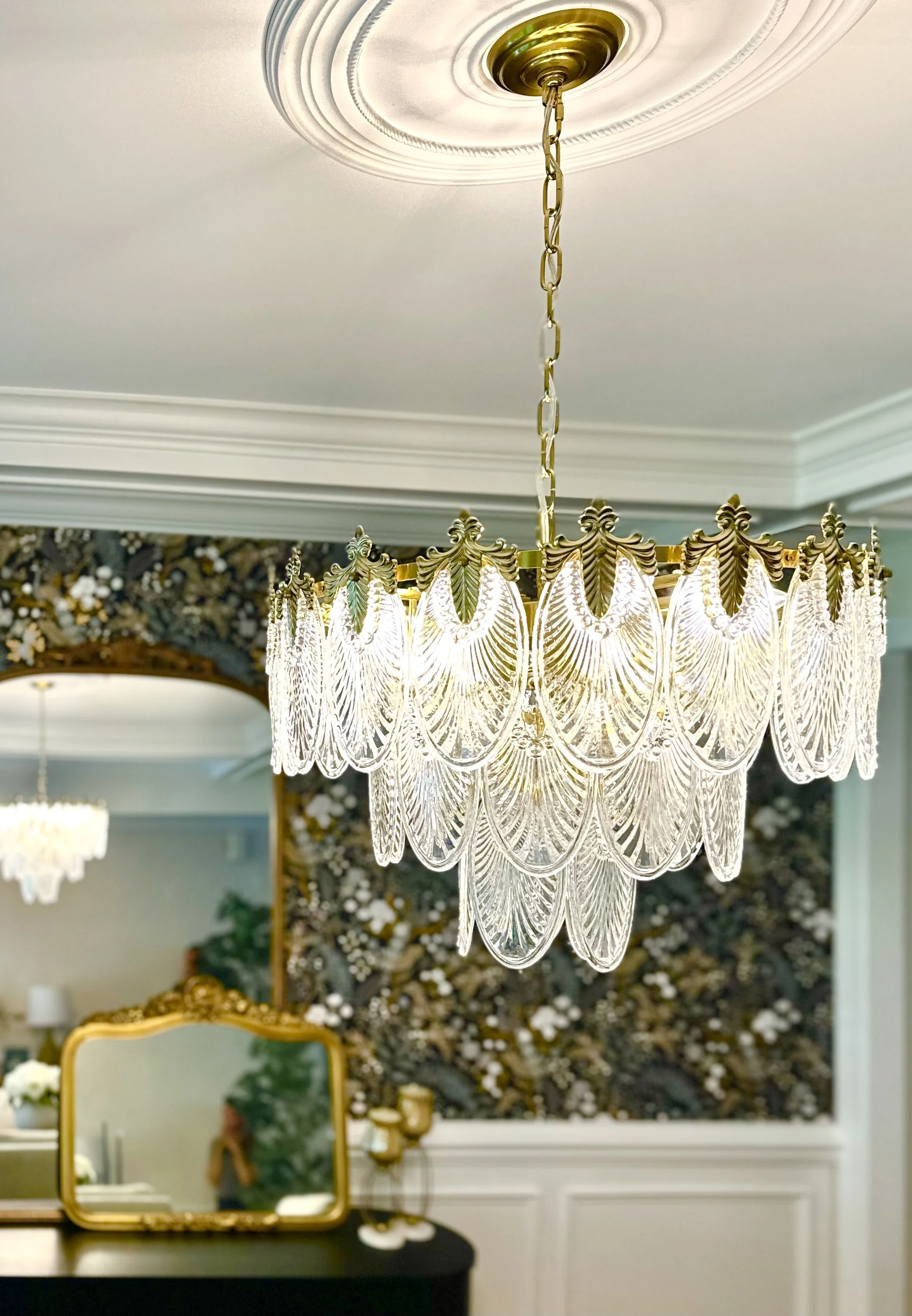 A decorative chandelier with ornate glass and gold accents hangs from a white ceiling in a room with floral wallpaper and a mirror reflecting part of the room.