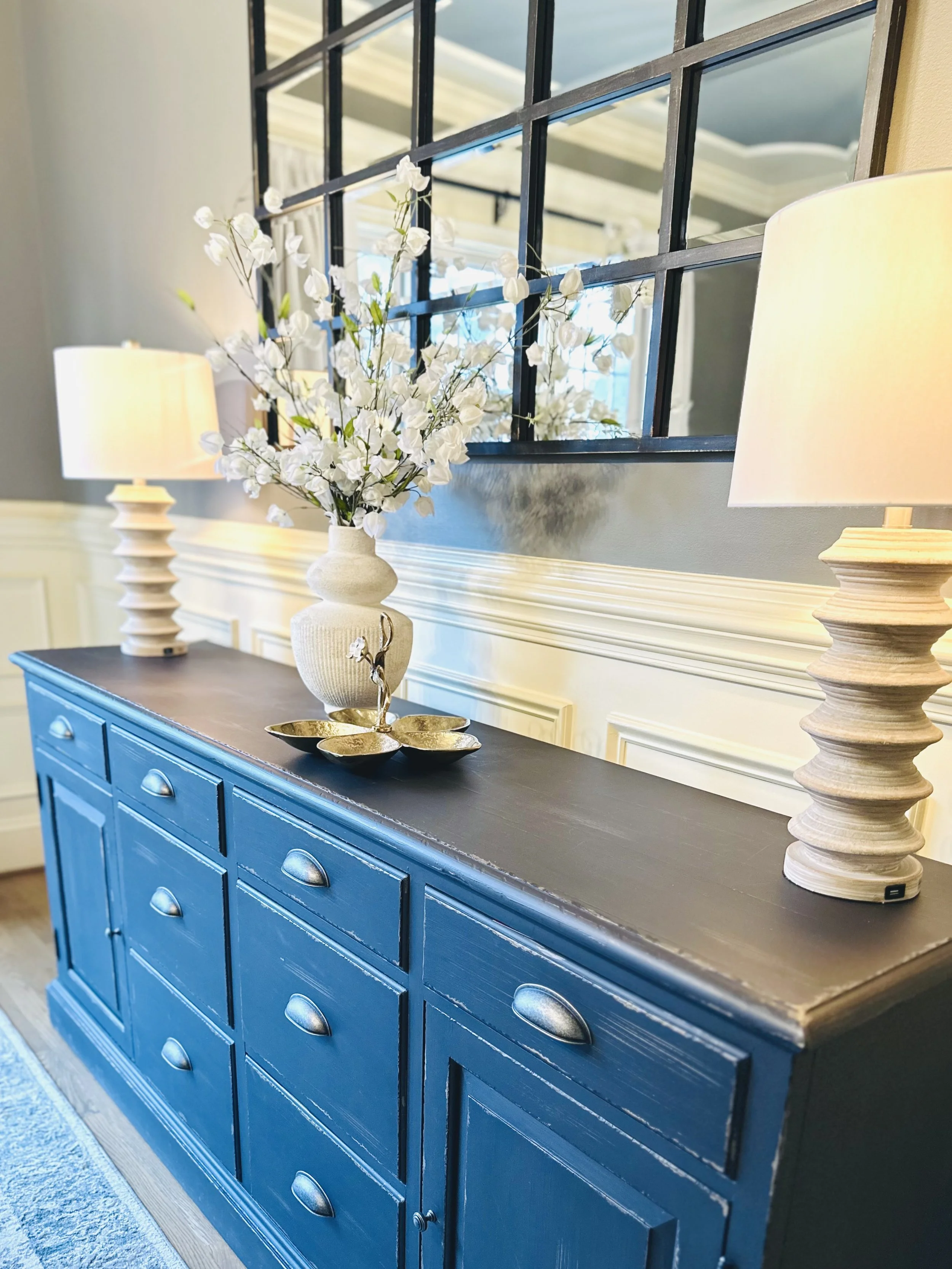 A blue sideboard with nine drawers, topped with a white vase containing white flowers, two table lamps, and three gold decorative bowls, with a mirror on the wall behind.