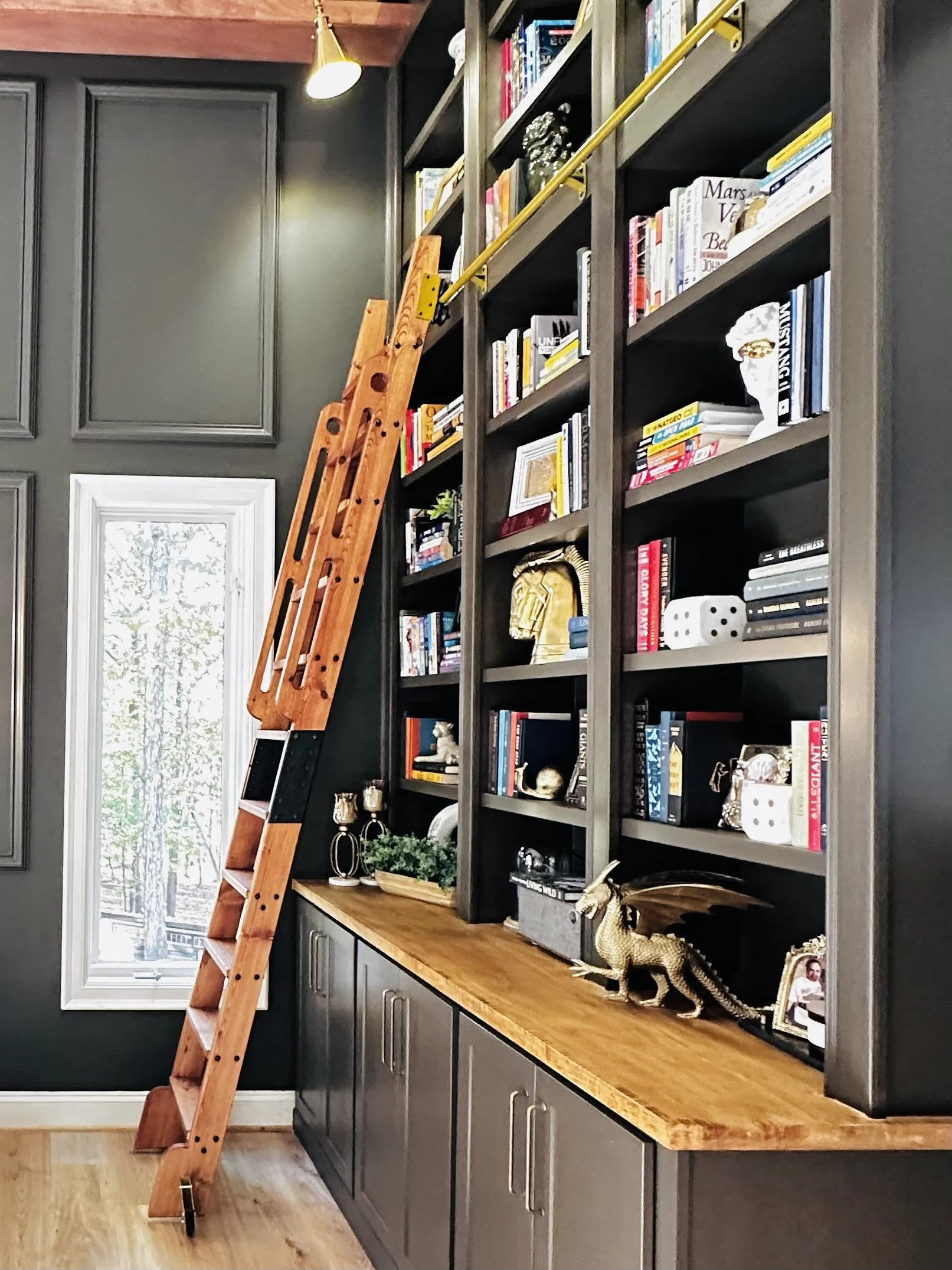 View of a corner bookshelf with decorative items and a wooden ladder leaning against it, in a room with dark gray walls and a window.